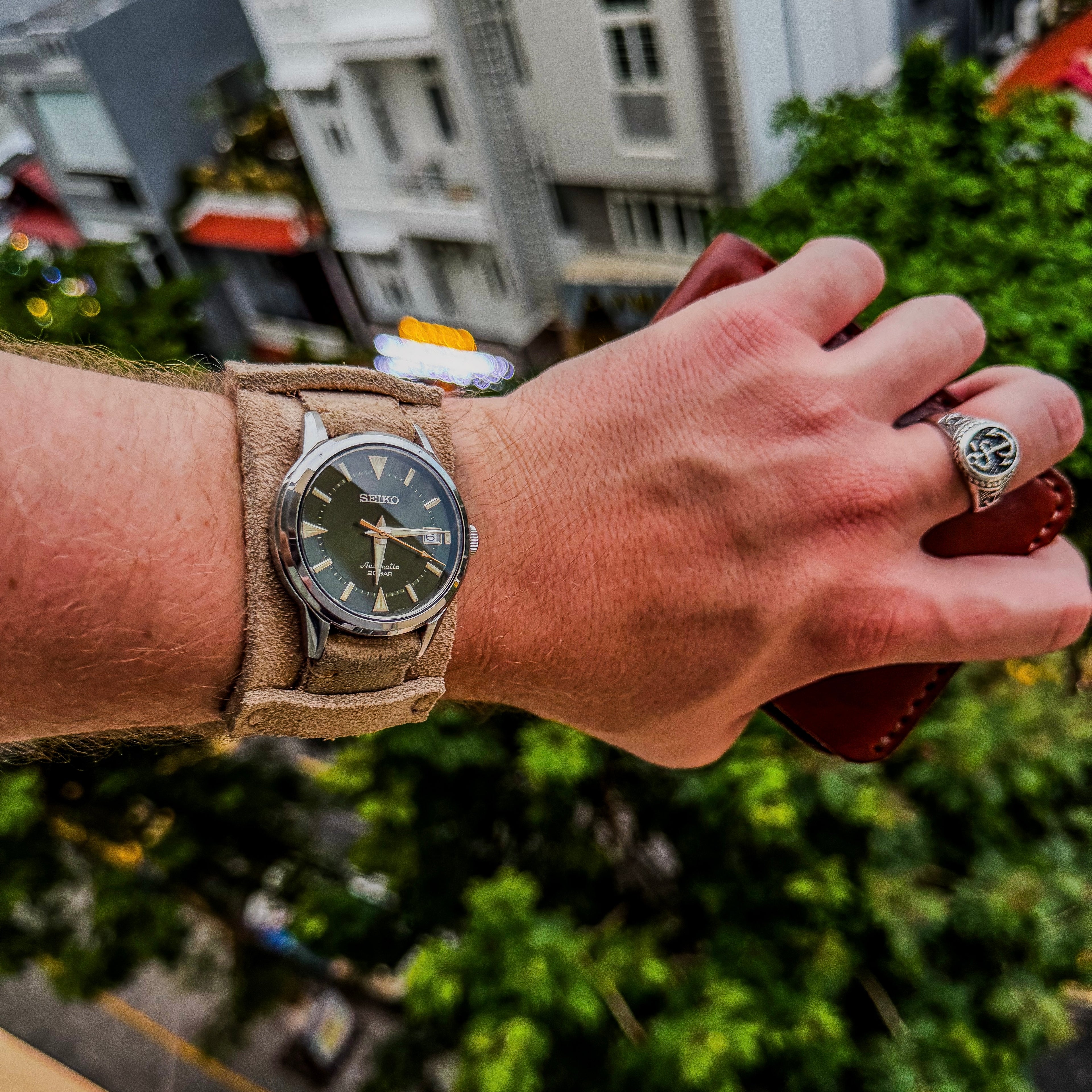 Close-up of a Seiko Alpinist watch on No. 53 Explorer Suede Watch Strap with bund pad, with a blurred cityscape of rooftops and trees in the background.