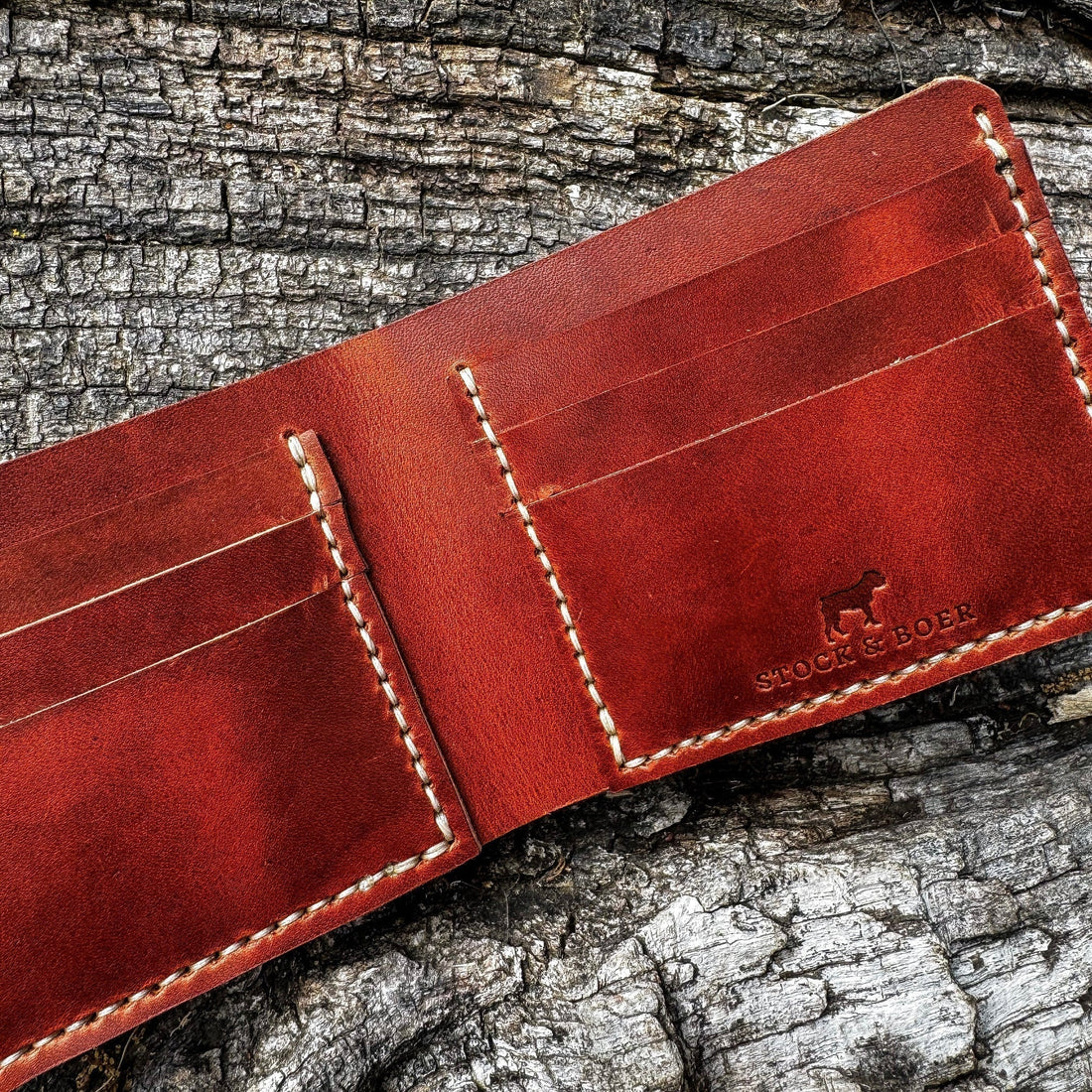 An interior view of an open, handcrafted full-grain leather bifold wallet featuring prominent cream hand-stitching and the embossed Stock & Boer logo, set against a rustic, weathered wood backdrop for the ultimate heritage carry guide.