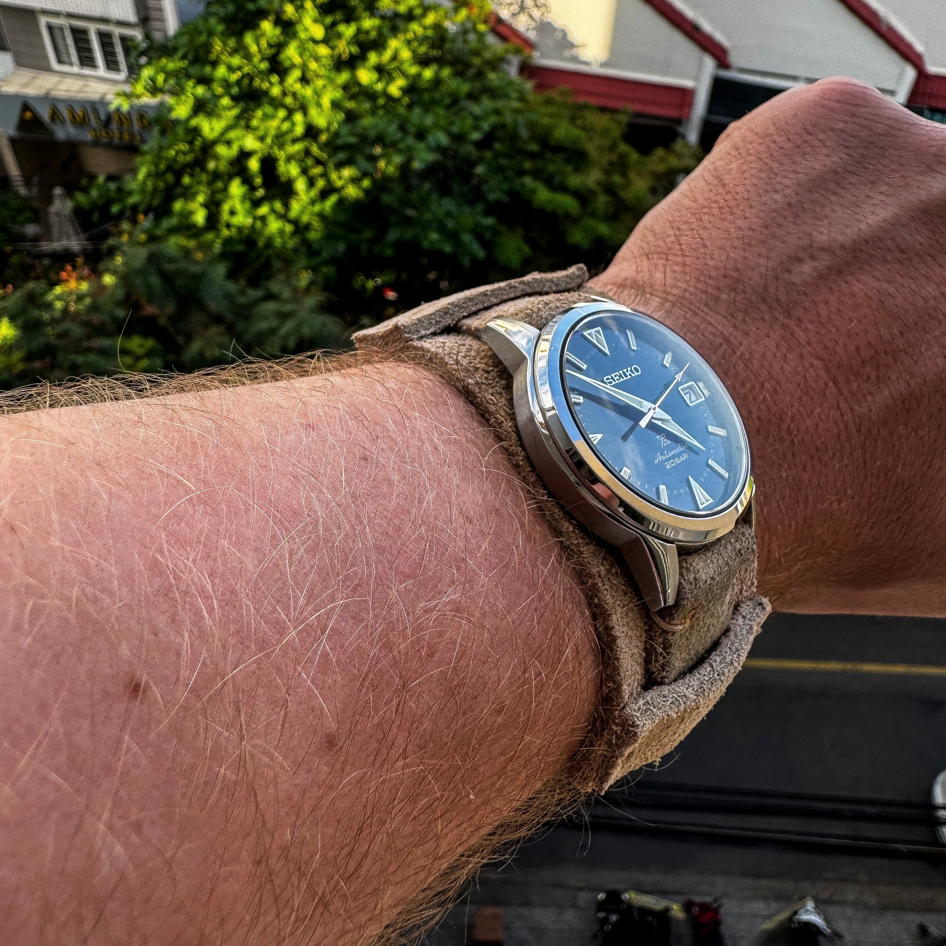 Wrist shot of a Seiko Alpinist watch on No. 53 Explorer Suede Watch Strap with bund pad, arm extended over a city street view with bright rooftops, trees, and a golden hour sky.