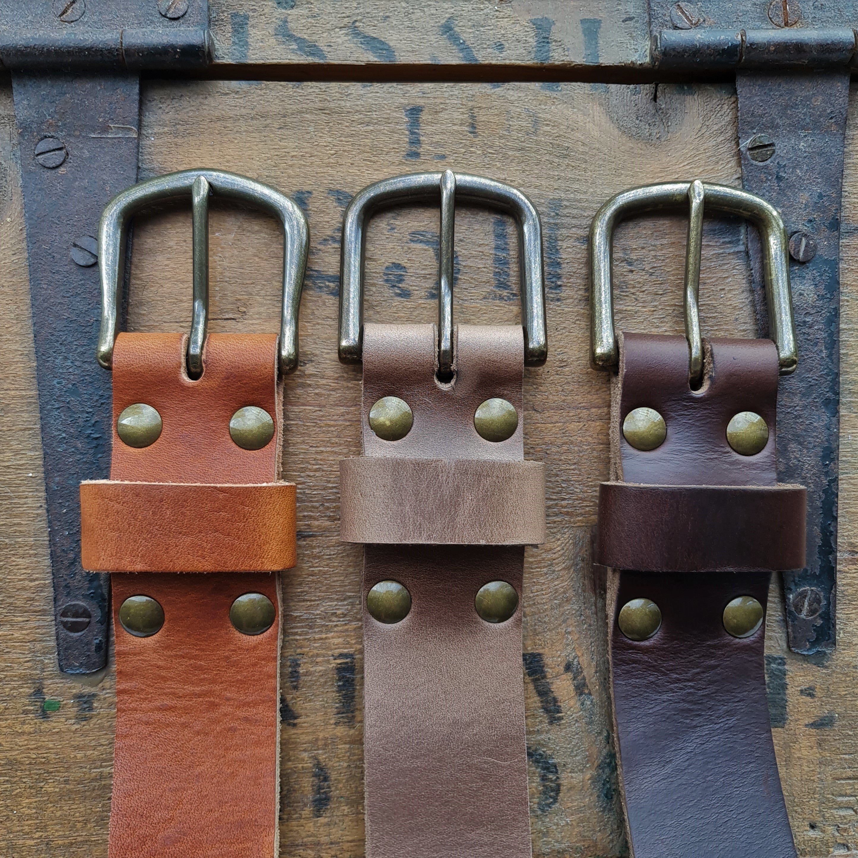 Three Horween leather belt buckles in natural Chromexcel, and brown and burgundy Chromexcel displayed on a vintage wooden background.