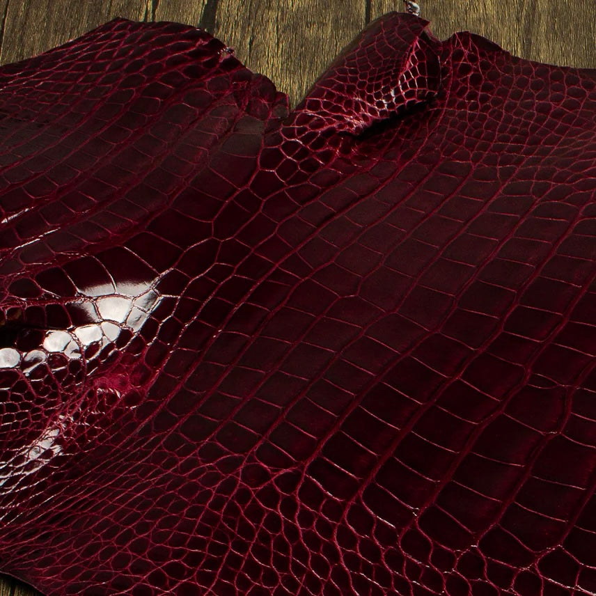 An overhead perspective of a premium Bull's Blood burgundy HCP Tannery alligator hide, displaying the natural, symmetrical scale structure and elegant finish of the exotic skin laid against a rustic wooden surface.