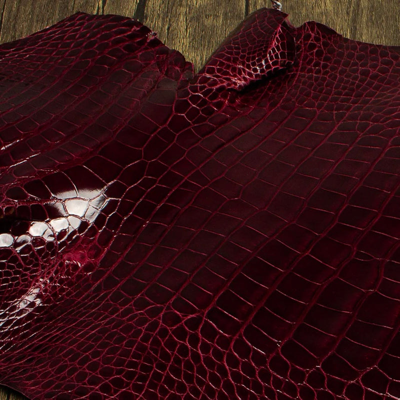 An overhead perspective of a premium Bull's Blood burgundy HCP Tannery alligator hide, displaying the natural, symmetrical scale structure and elegant finish of the exotic skin laid against a rustic wooden surface.