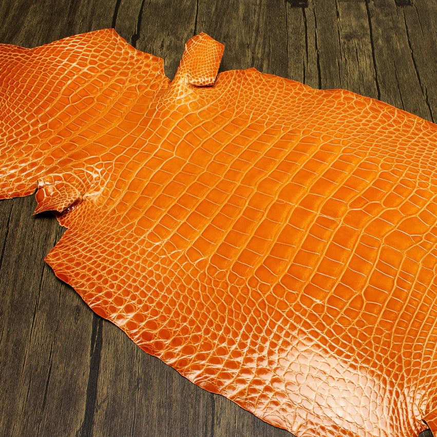 A high-angle, detailed shot of a vibrant mandarin orange HCP Tenery alligator hide, showcasing its symmetrical scale pattern and luxurious finish against a rustic, weathered wood background.