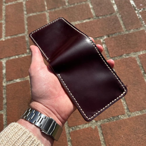 A hand holding the No. 1 WRANGLER Leather Card Holder in a closed position, highlighting the deep, polished burgundy finish of the Horween Chromexcel leather. The card holder is held against a background of reddish-brown brick paving.