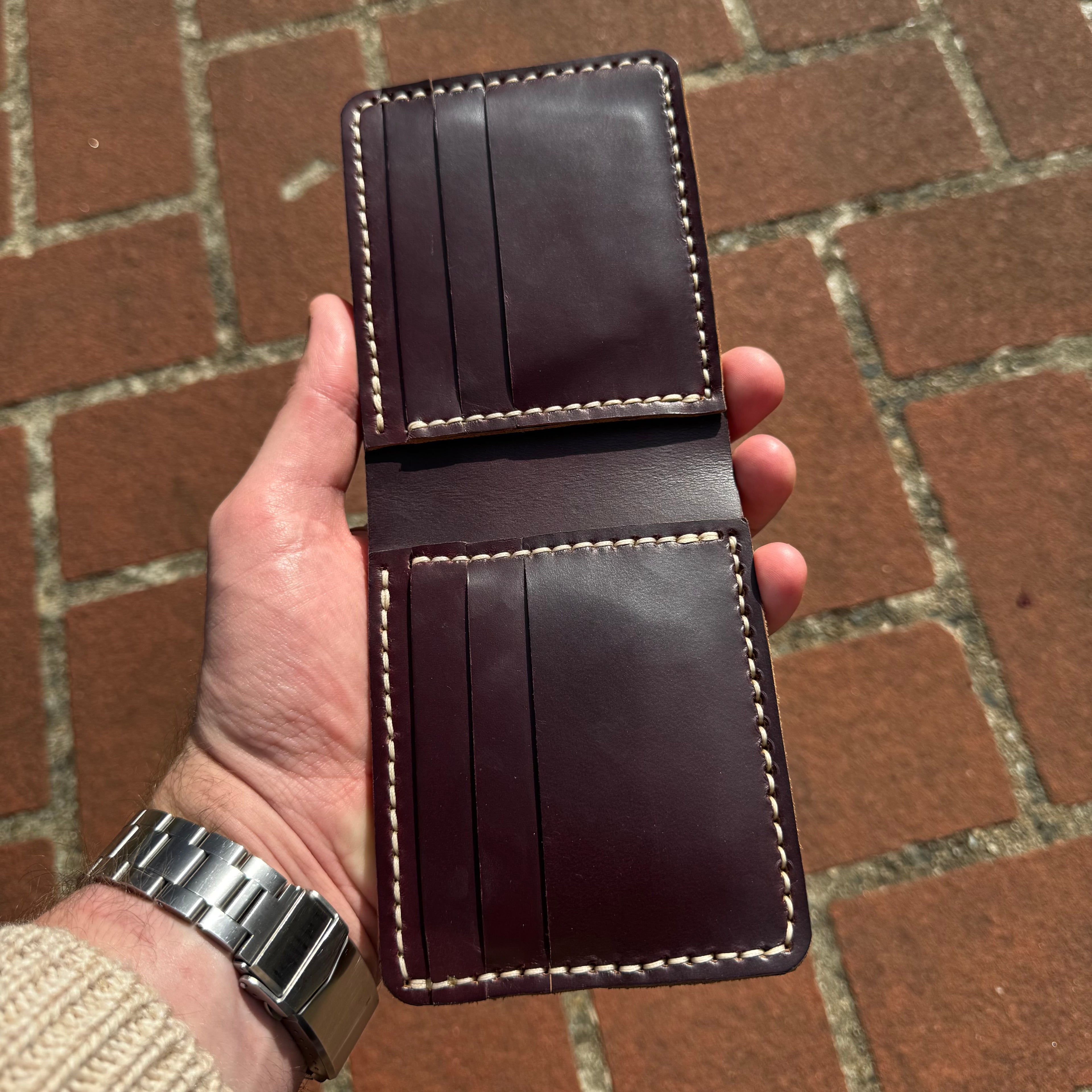 A hand holding the No. 1 WRANGLER Leather Card Holder open, revealing the internal card slots and contrast stitching. The card holder is made of burgundy Horween Chromexcel leather and is positioned over reddish-brown brick paving.