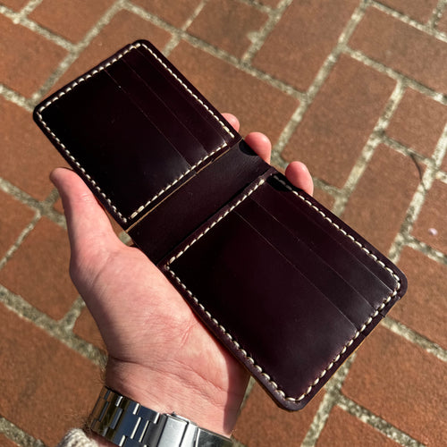 A hand holding the No. 1 WRANGLER Leather Card Holder in an open position, showcasing the interior slots and detailed stitching. The card holder is crafted from rich, burgundy Horween Chromexcel leather and is held against a background of reddish-brown brick paving.