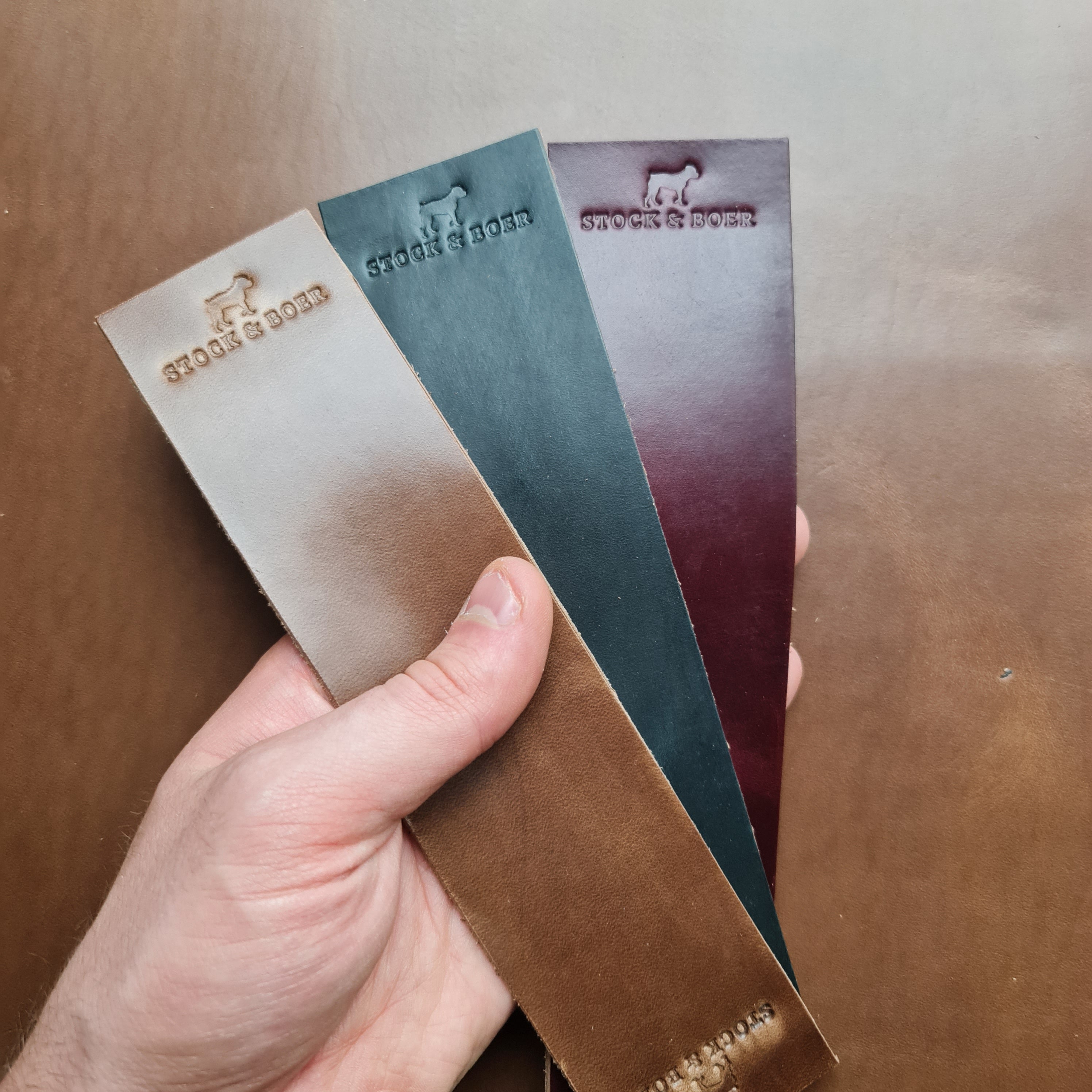 A hand holding three vertical No. 11 RAILMAN Bookmarks made from Horween Chromexcel leather, each stamped with the "STOCK & BOER" logo. The bookmarks are displayed in three distinct colors: natural, burgundy, and navy. They are held against a blurred brown leather background.