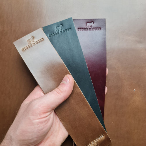 A hand holding three vertical No. 11 RAILMAN Bookmarks made from Horween Chromexcel leather, each stamped with the "STOCK & BOER" logo. The bookmarks are displayed in three distinct colors: natural, burgundy, and navy. They are held against a blurred brown leather background.