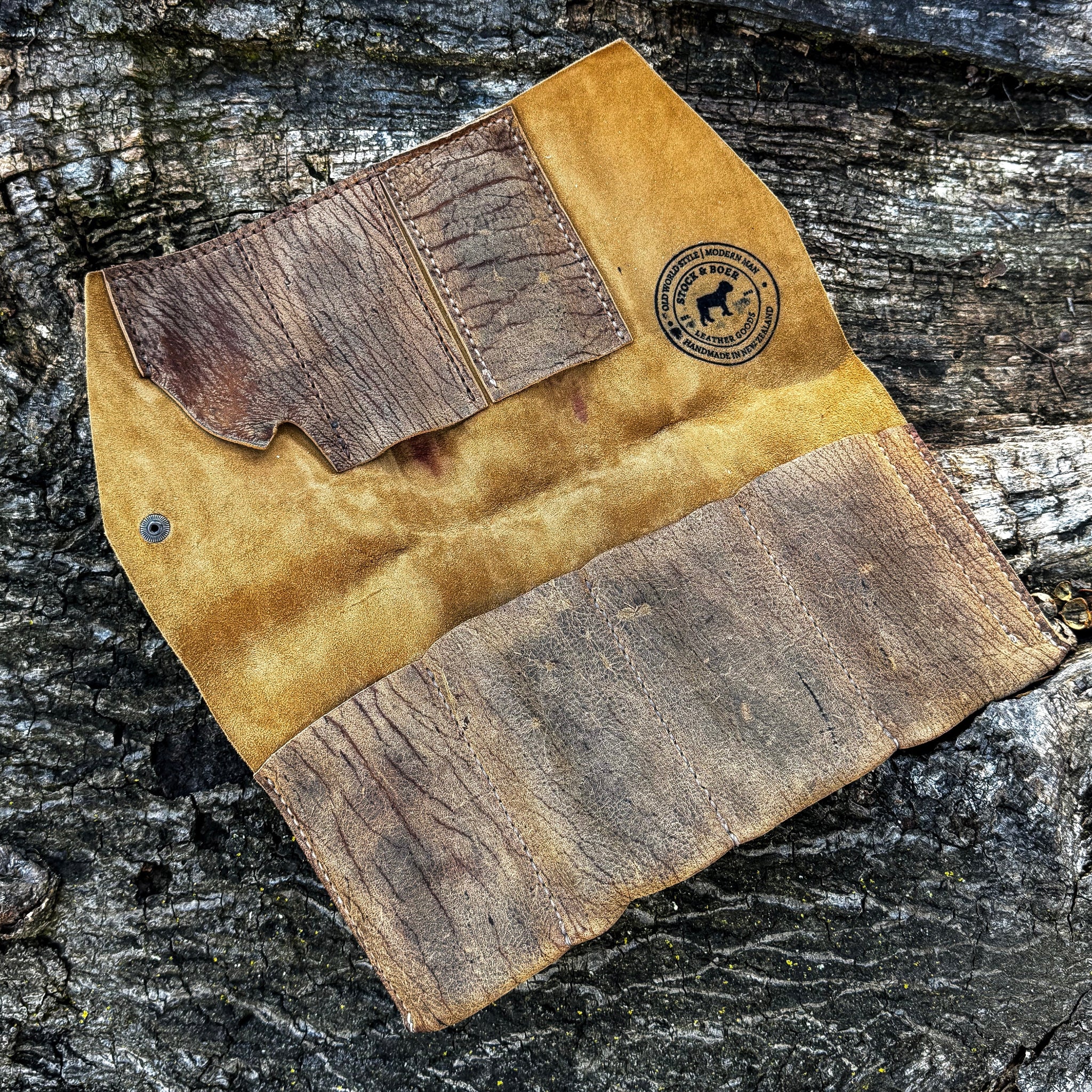 The interior view of an open No. 13 Plunderer Leather Watch Roll crafted from Baobab Wild African Kudu leather. The image highlights the soft golden suede inner lining, a circular Stock & Boer stamped logo, internal storage pockets, and the rugged, naturally scarred leather of the flaps, all laid flat against a textured tree bark background.