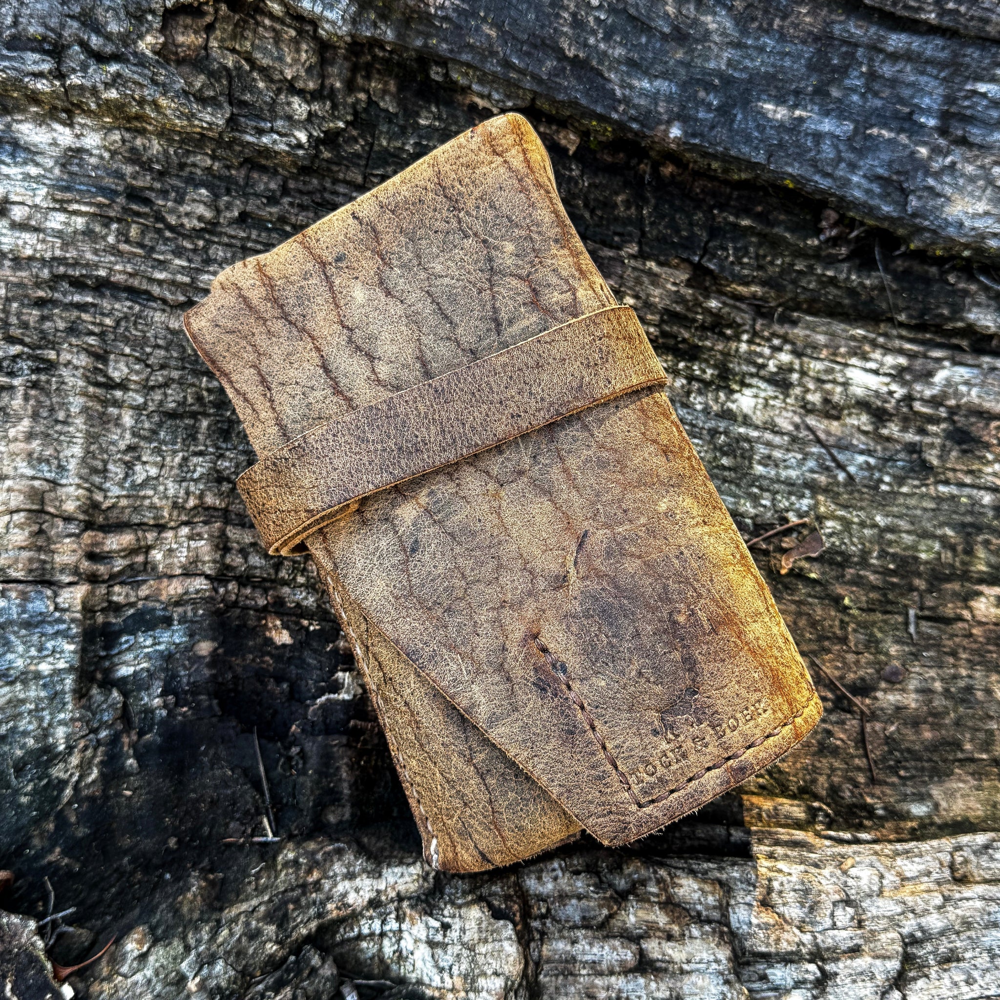 A rolled and secured No. 13 Plunderer Leather Watch Roll crafted from heavily textured Baobab Wild African Kudu leather. The watch roll rests on a piece of rustic, weathered tree bark, showcasing the prominent natural scarring and rugged grain of the hide.