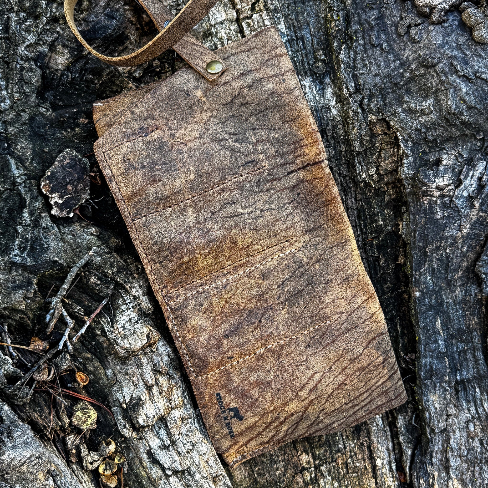 The exterior view of the No. 13 Plunderer Leather Watch Roll laid flat, crafted from heavily textured Baobab Wild African Kudu leather. The image showcases the natural hide striations, vertical stitching lines indicating the internal pockets, an attached wrap strap with a metal stud, and a subtle Stock & Boer logo stamped in the corner, set against a rugged tree bark background.
