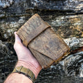 A hand holding the No. 13 Plunderer Leather Watch Roll crafted from highly textured Baobab Wild African Kudu leather, featuring natural scars, hand-stitched details, and a wrap-around strap, set against a rugged tree bark background.