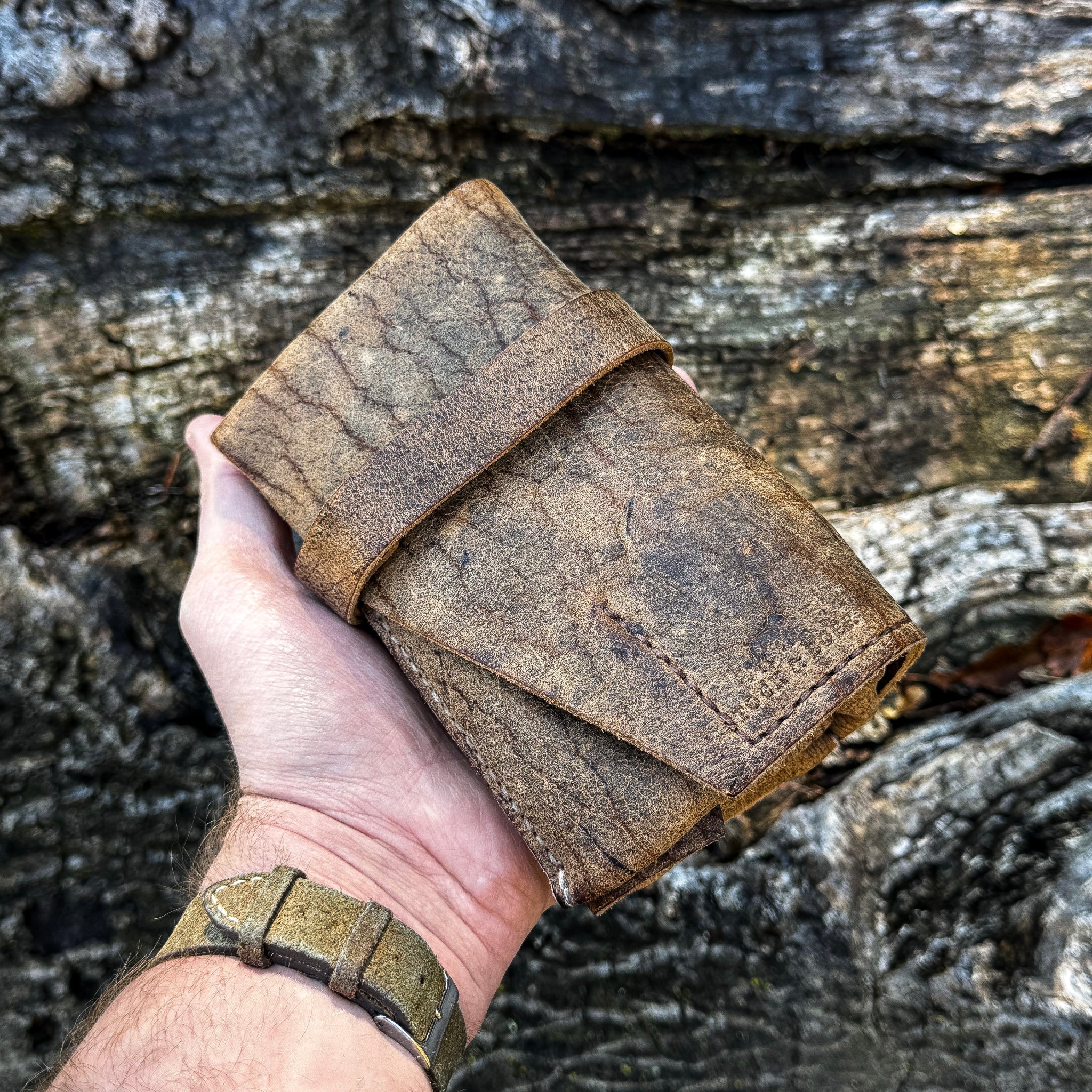 A hand holding the No. 13 Plunderer Leather Watch Roll crafted from highly textured Baobab Wild African Kudu leather, featuring natural scars, hand-stitched details, and a wrap-around strap, set against a rugged tree bark background.