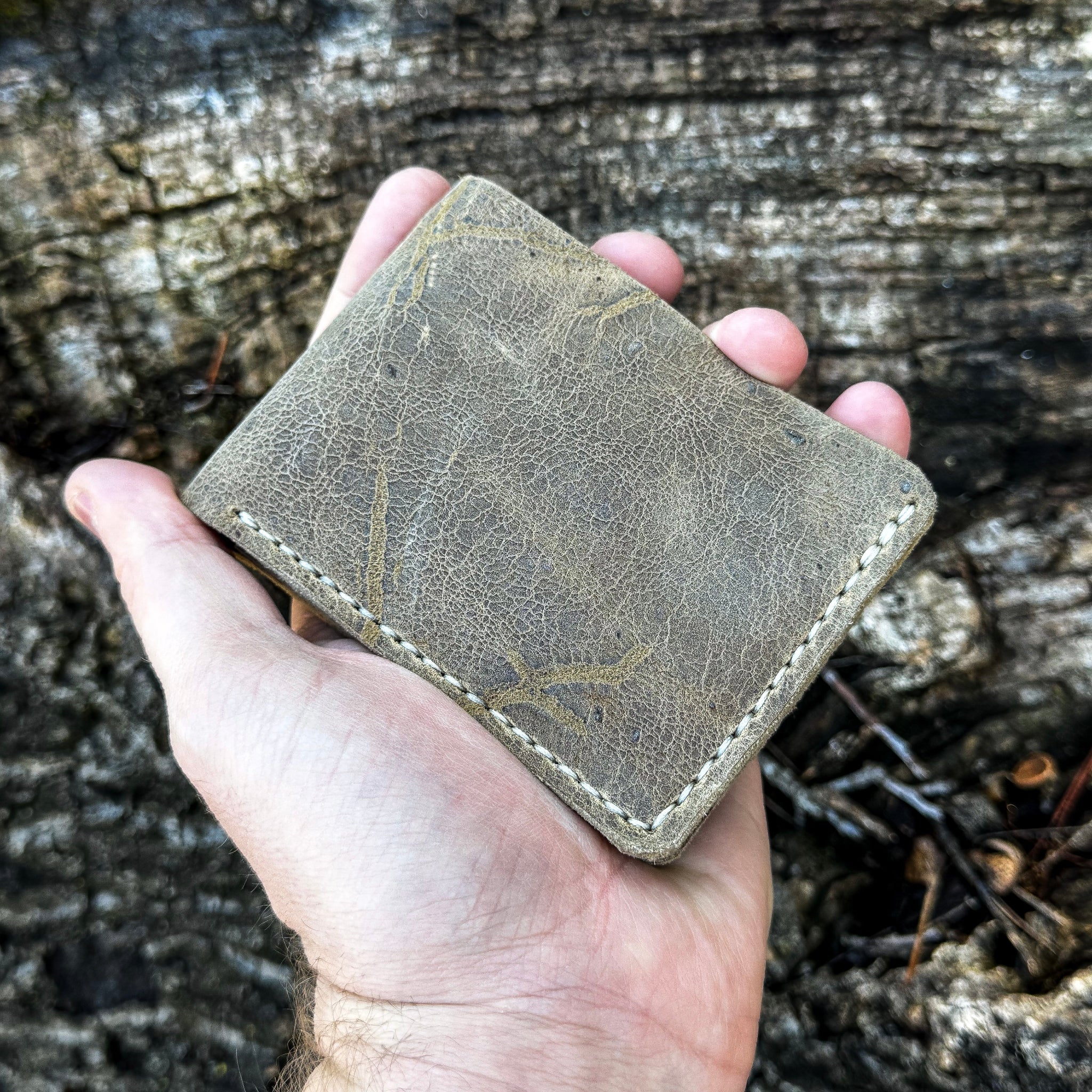 A hand cradling the closed handcrafted No. 1652 JAGTER field bifold wallet in Desert color Wild African Kudu leather, showcasing the rugged natural hide texture and perimeter hand-stitching.