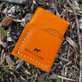 Overhead view of the No. 1835 Trekker Front Pocket Wallet crafted from iconic Horween MLB baseball leather, featuring the distinctive bright orange hide, contrast stitching, and Stock & Boer branding, set against a rugged, natural forest floor background.