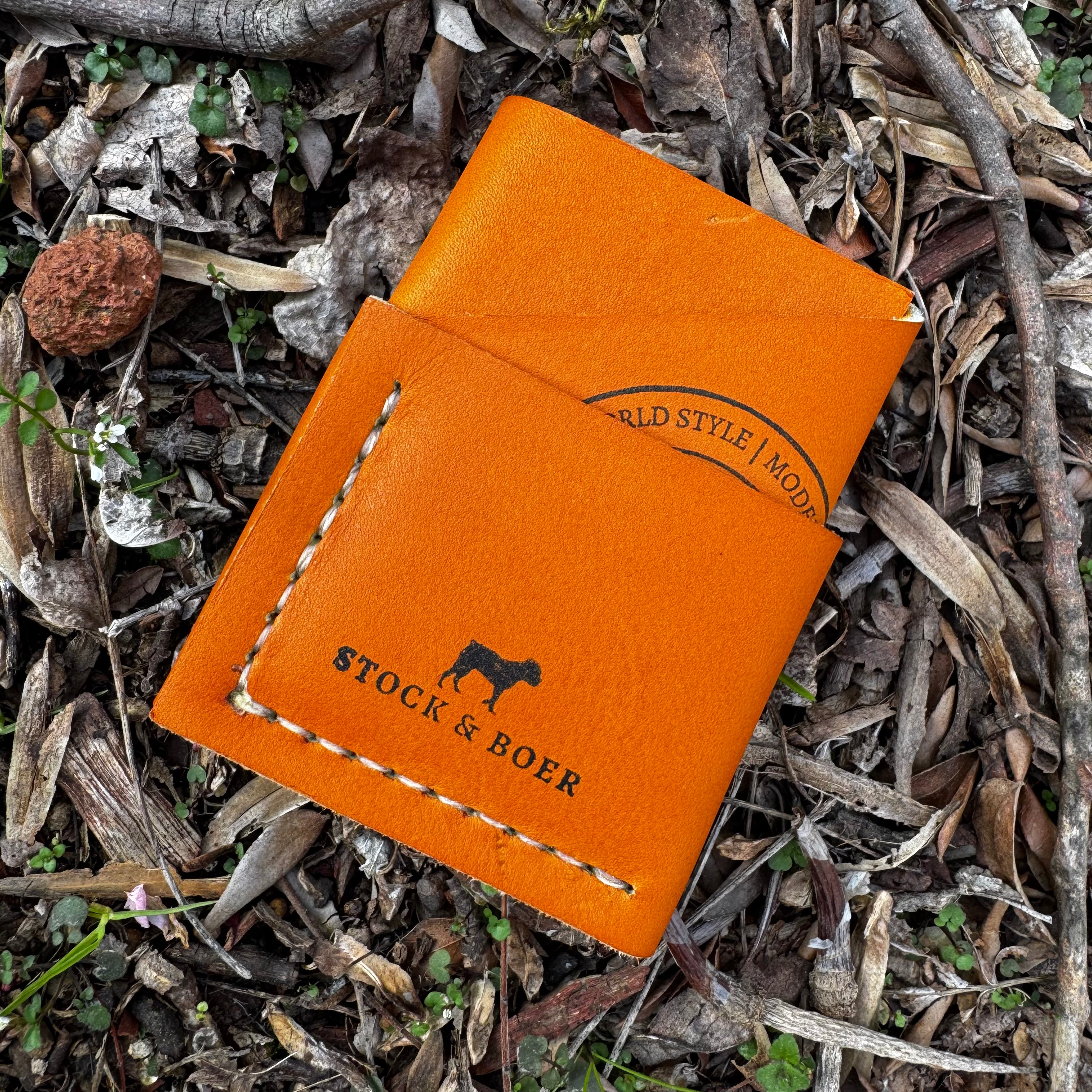 Overhead view of the No. 1835 Trekker Front Pocket Wallet crafted from iconic Horween MLB baseball leather, featuring the distinctive bright orange hide, contrast stitching, and Stock & Boer branding, set against a rugged, natural forest floor background.