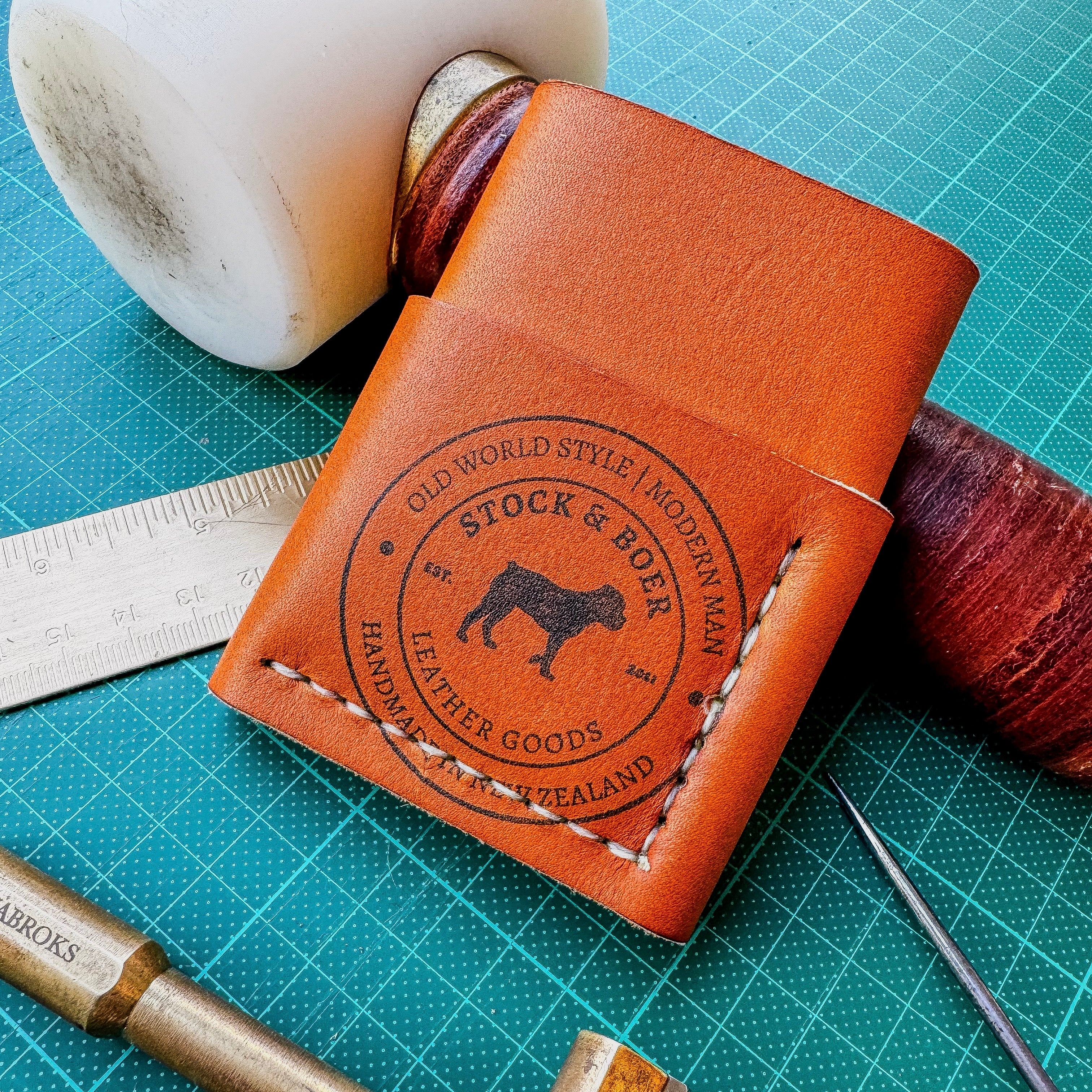 Detailed view of the No. 1835 Trekker Front Pocket Wallet in Horween MLB baseball leather, showcasing the ink stamp Stock & Boer maker's mark and hand-stitched details resting on a green professional cutting mat surrounded by leatherworking tools.