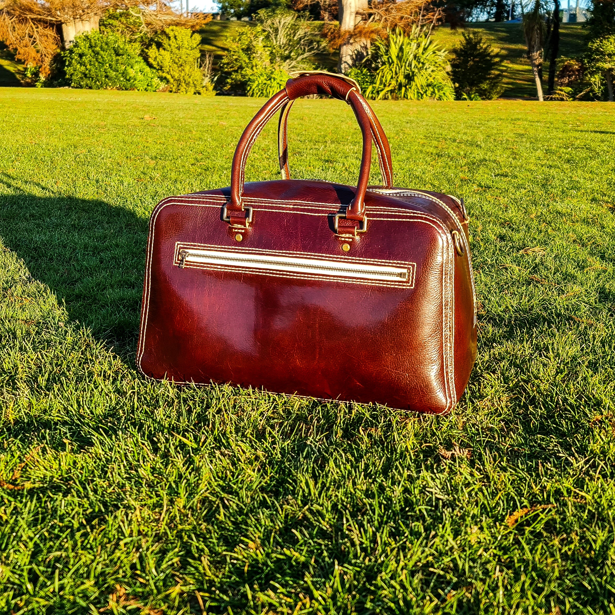 An angled lifestyle shot of the No. 1963 VOYAGER Travel Bag in a glossy Tuscan Brown leather, resting on bright green grass. The bag features prominent white hand-stitching, rolled leather top handles, and a horizontal front zippered pocket with brass hardware, illuminated by warm golden-hour sunlight casting a shadow to the left against a softly blurred background of park trees.