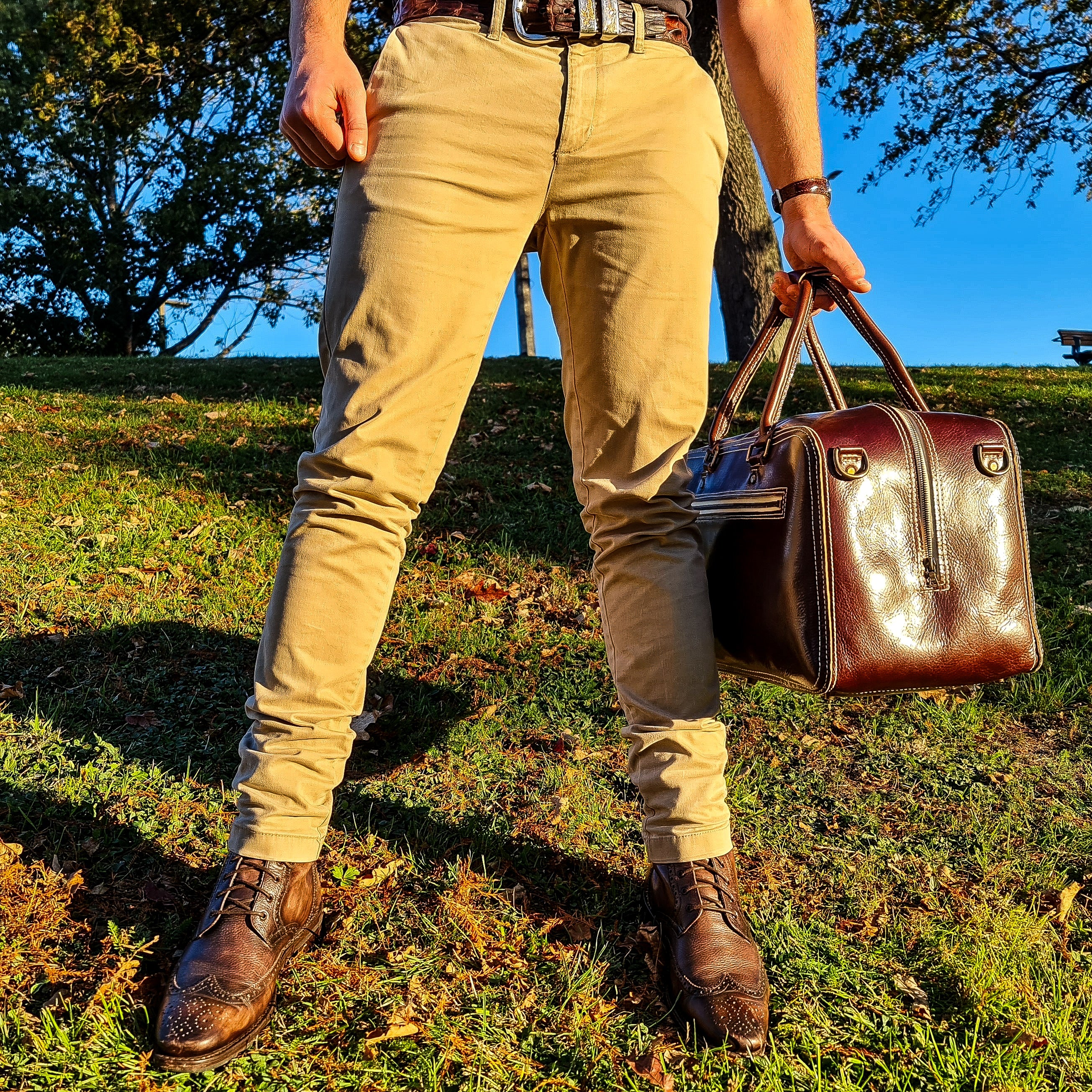 A lifestyle shot showing a person's lower half from the front as they carry the No. 1963 VOYAGER Travel Bag by its rolled top handles. The glossy Tuscan Brown leather bag is angled to reveal both the front zippered pocket and the side vertical zipper, featuring prominent white hand-stitching and brass hardware. The person is styled in tan pants, a brown leather belt, and brown leather brogue boots, standing on a grassy hill in a park illuminated by warm golden-hour sunlight, casting long shadows with trees 
