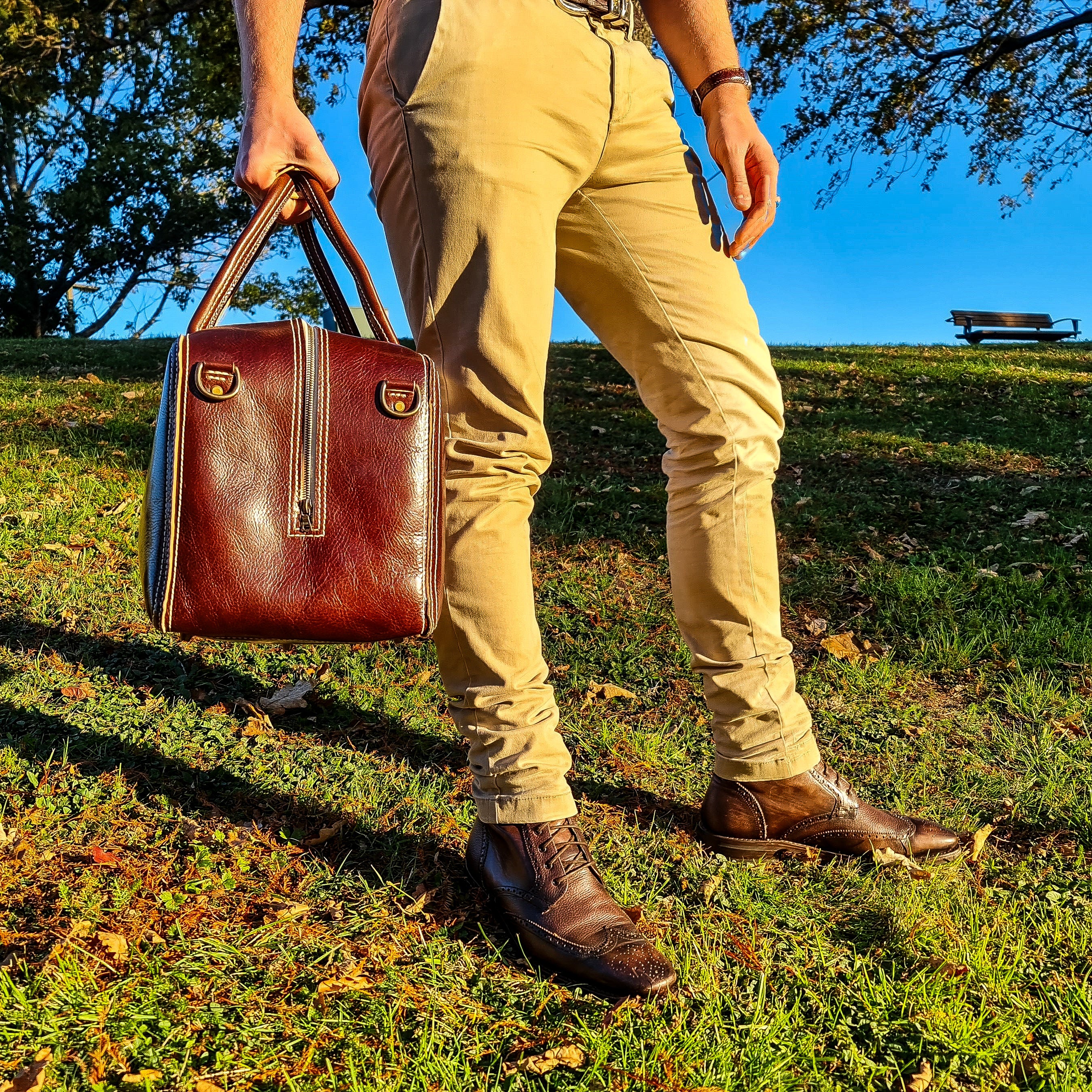 A lifestyle shot showing a person's lower half as they carry the No. 1963 VOYAGER Travel Bag by its rolled top handles. The side profile of the glossy Tuscan Brown leather bag is visible, showcasing prominent white hand-stitching, a vertical side zipper, and brass D-rings. The person is styled in tan pants and brown leather brogue boots, standing on a grassy hill in a park illuminated by warm golden-hour sunlight, with trees and a park bench in the background under a clear blue sky.