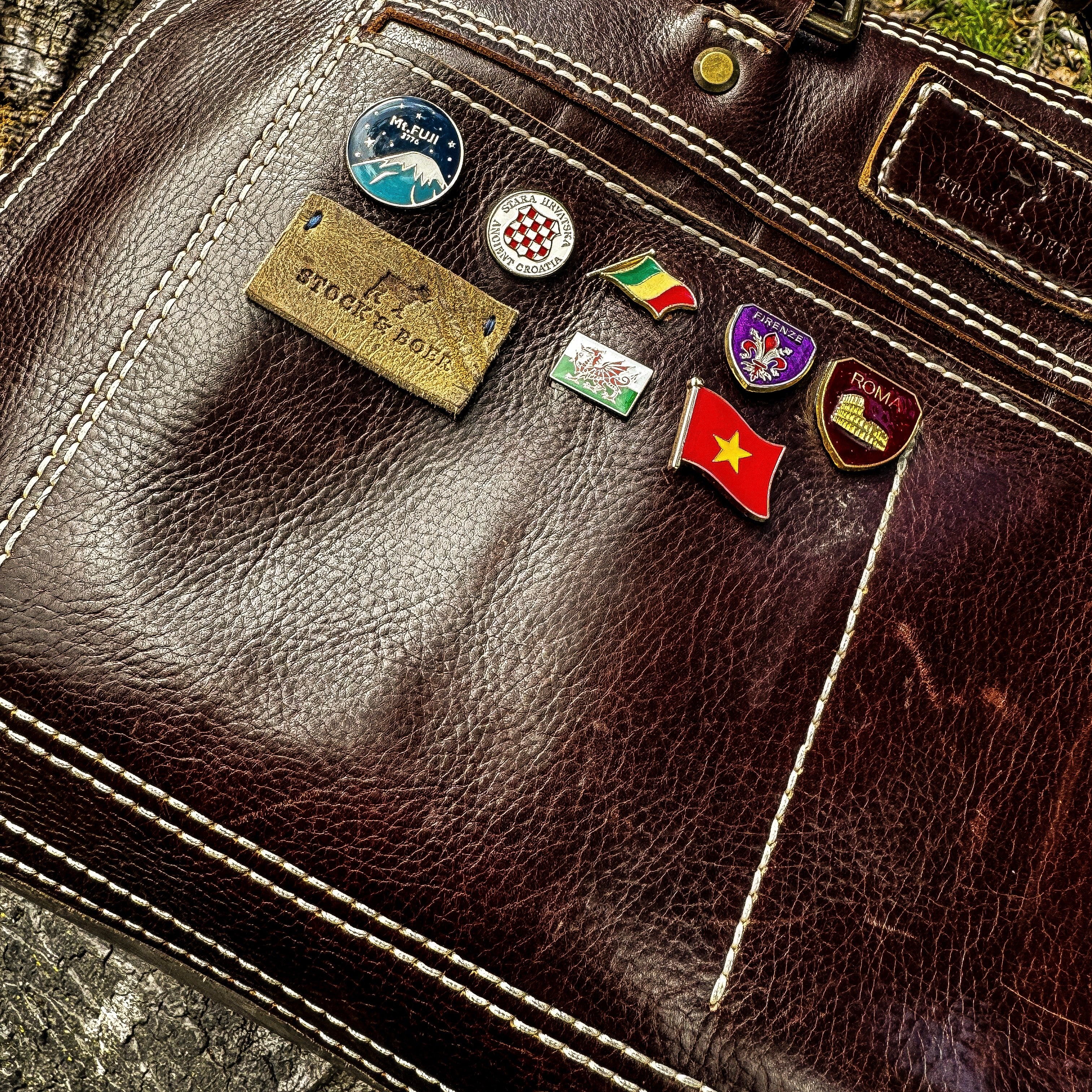 A close-up shot focusing on the front slip pocket of the No. 1963 VOYAGER Travel Bag in a glossy Tuscan Brown leather. The richly textured surface is personalized with a collection of colorful enamel travel pins—including flags and city crests—alongside a small rectangular leather logo patch. The image highlights the bag's prominent white hand-stitching and a glimpse of the brass hardware, set against a rough, weathered wooden background.