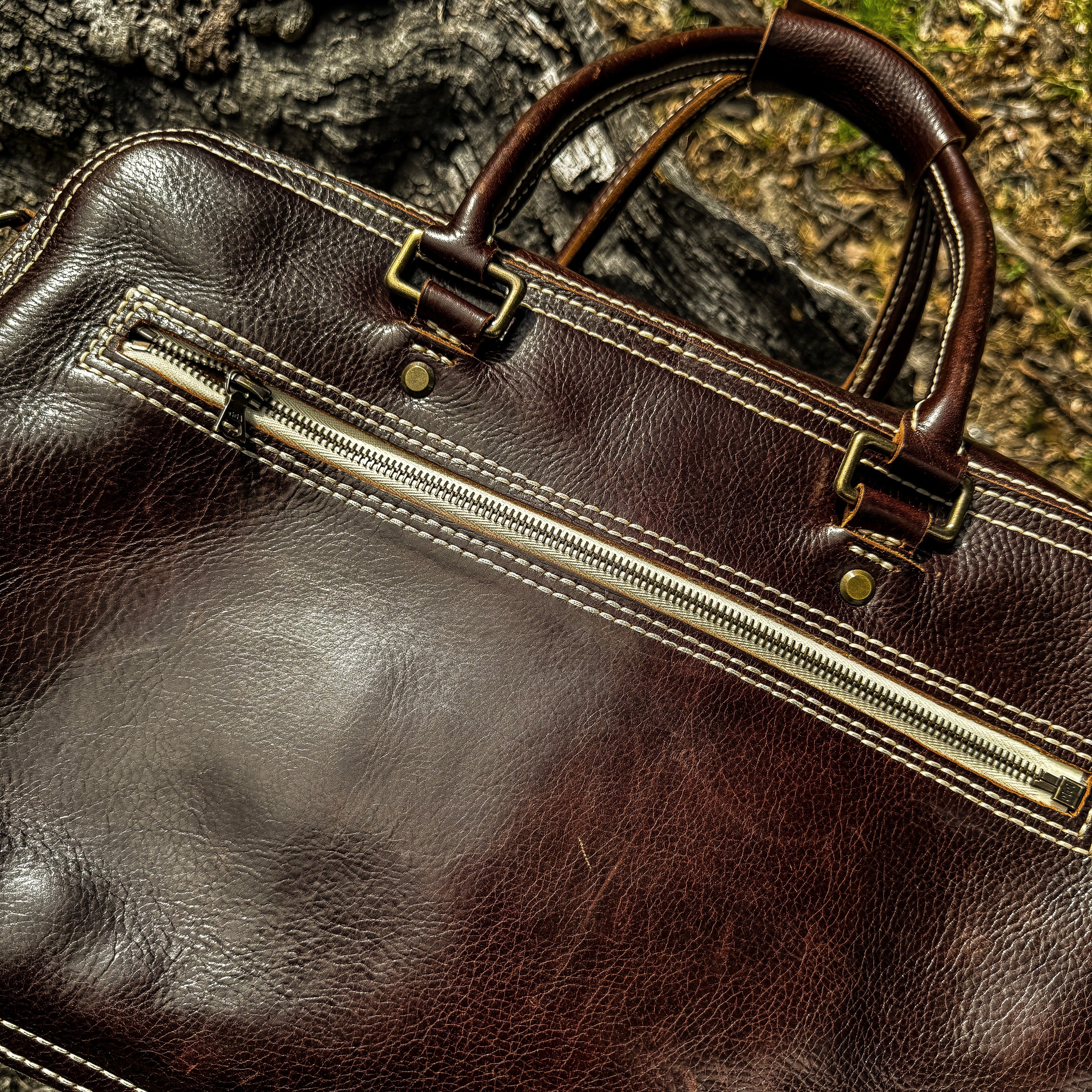 A close-up lifestyle shot of the No. 1963 VOYAGER Travel Bag in a glossy Tuscan Brown leather, resting on a highly textured, weathered tree trunk. The image focuses closely on the bag's detailed craftsmanship, highlighting the horizontal front zippered pocket, prominent white hand-stitching, brass rivets, and the base of the rolled leather top handles set against a natural, rugged outdoor background.