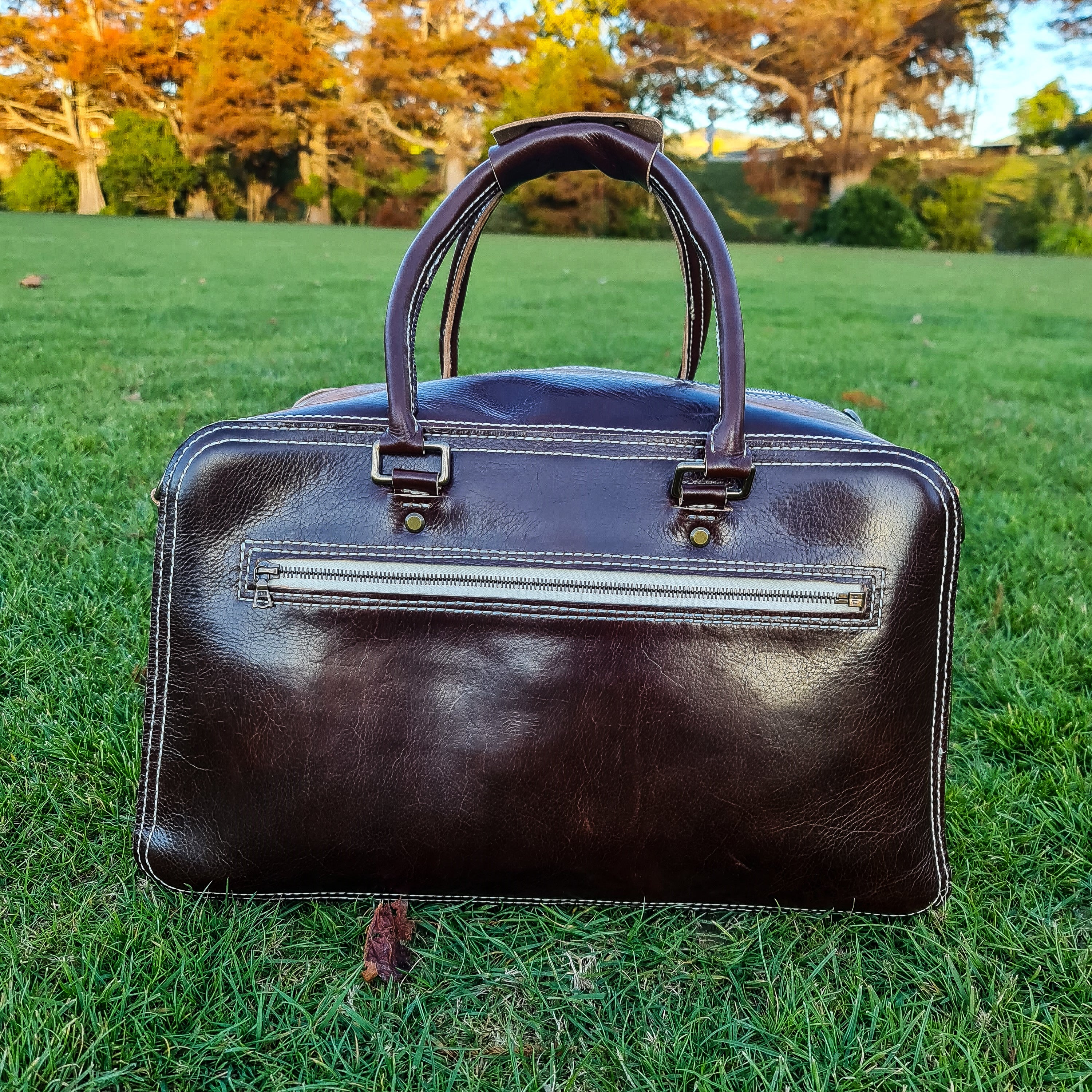 A lifestyle front-view shot of the No. 1963 VOYAGER Travel Bag in a rich Tuscan Brown leather, resting on bright green grass. The bag features prominent white hand-stitching, sturdy rolled leather top handles, and a horizontal front zippered pocket with brass hardware, set against a softly blurred background of park trees with autumnal foliage.