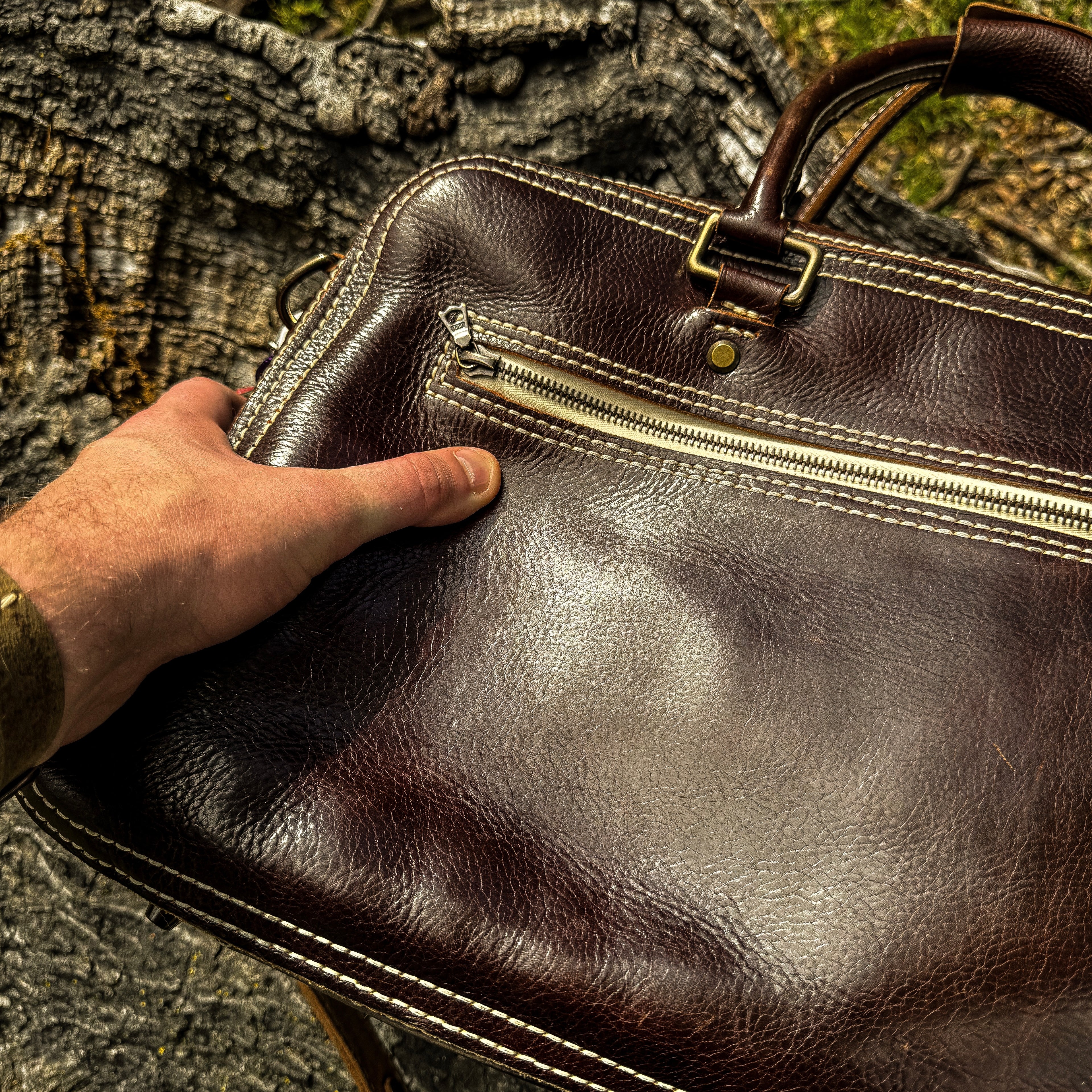 A close-up lifestyle shot showing a person's hand firmly gripping the side edge of the No. 1963 VOYAGER Travel Bag in a glossy Tuscan Brown leather. The bag rests against a highly textured, weathered tree trunk, emphasizing the rich, natural grain of the hide. The image highlights the bag's prominent white hand-stitching along the edges, the horizontal front zippered pocket, brass hardware, and the base of the rolled leather top handle.