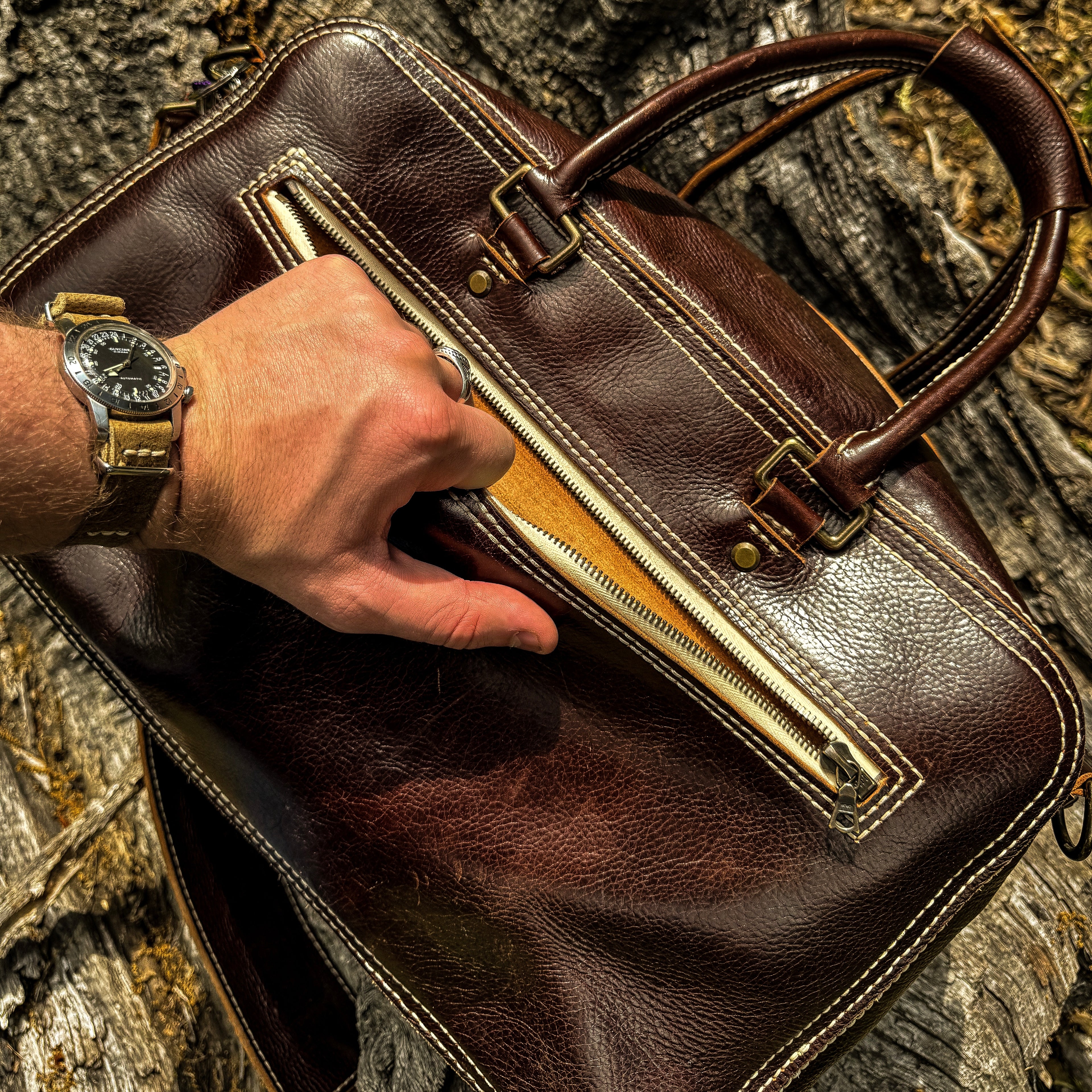 A close-up lifestyle shot showing a hand reaching into the partially unzipped front pocket of the No. 1963 VOYAGER Travel Bag in a glossy Tuscan Brown leather. The person, wearing a watch with a tan suede strap on their wrist, is interacting with the bag as it rests on a highly textured, weathered tree trunk. The image highlights the bag's prominent white hand-stitching, brass hardware, and the raw leather interior of the zippered pocket.
