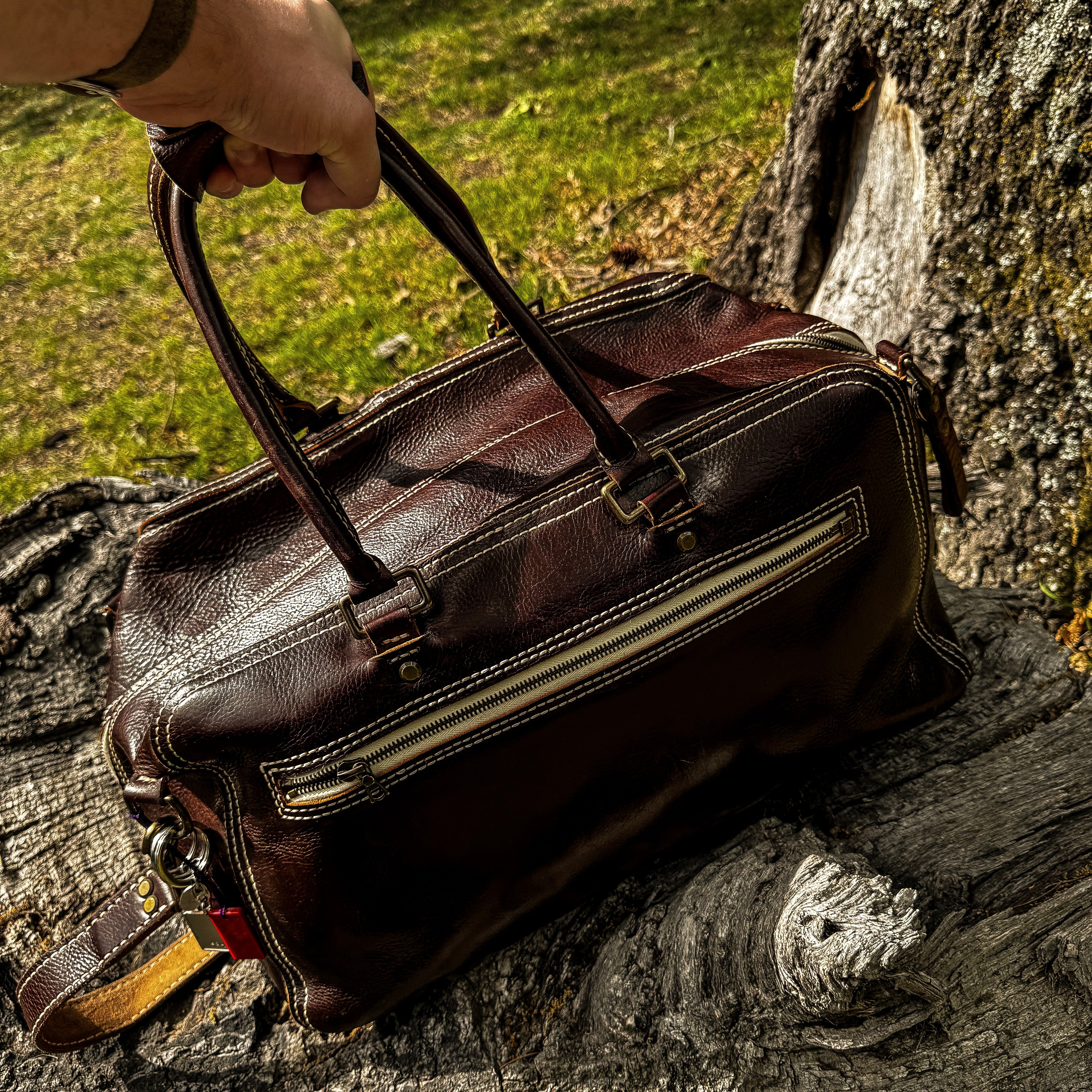 A close-up lifestyle shot showing a hand holding the No. 1963 VOYAGER Travel Bag by its rolled top handles. The rich Tuscan Brown leather bag rests partially on a highly textured, weathered tree trunk. The image highlights the bag's prominent white hand-stitching, horizontal front zippered pocket, brass hardware, and attached shoulder strap, set against a background of green grass and a tree base under bright sunlight.