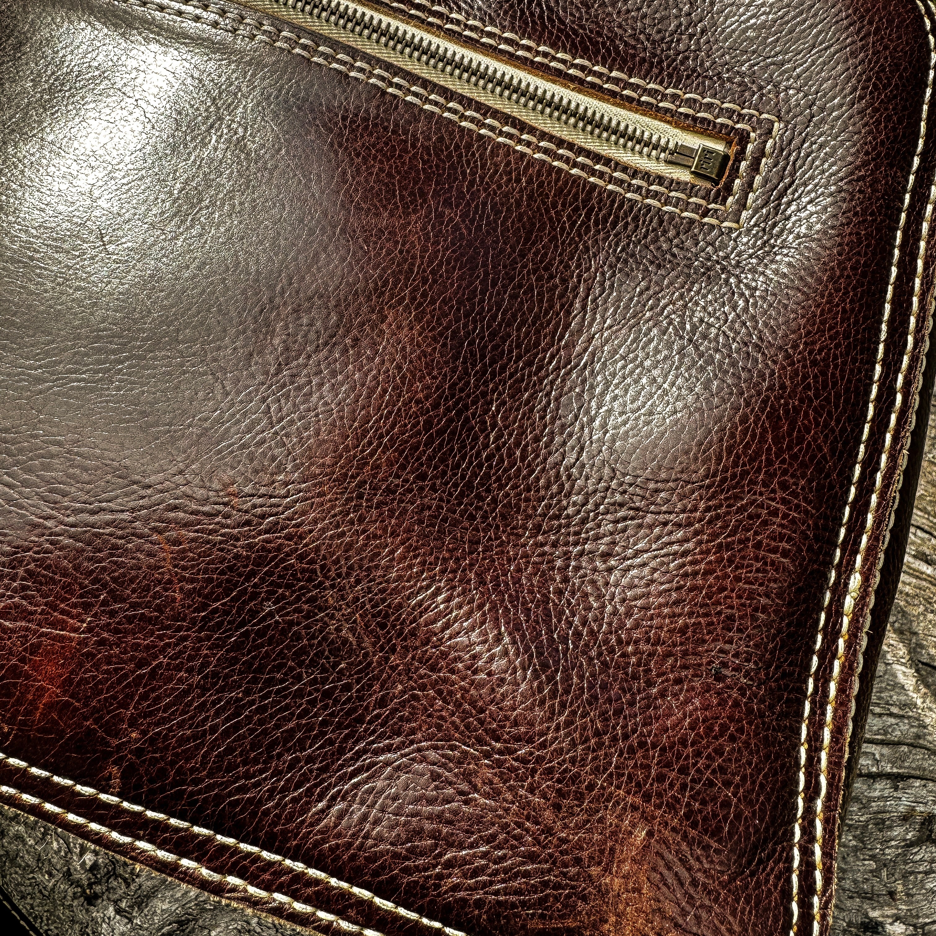 A macro close-up shot focusing on the exquisite texture of the No. 1963 VOYAGER Travel Bag in a glossy Tuscan Brown leather. The image details the rich, grainy surface and natural patina of the hide, accented by meticulous white hand-stitching along the bottom edge and framing a section of a metal zipper, set against a rough, weathered wooden background.