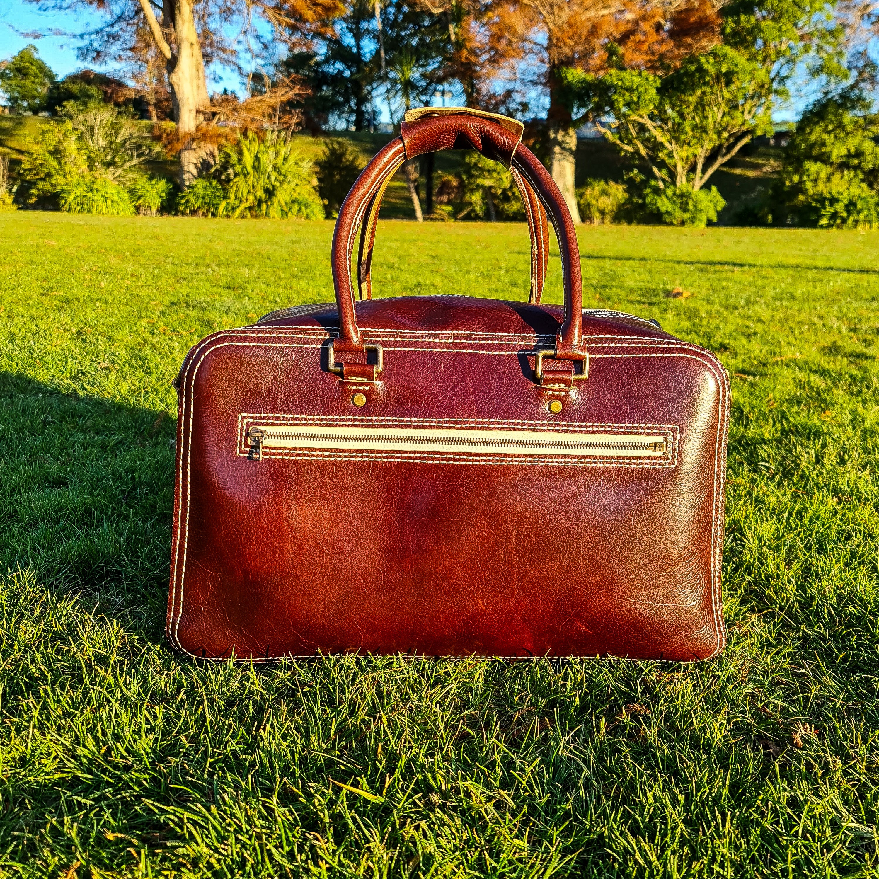 A lifestyle shot of the No. 1963 VOYAGER Travel Bag in a rich Tuscan Brown leather, resting on a bright green lawn. The bag features prominent white hand-stitching, sturdy rolled leather top handles, and a horizontal front zippered pocket with brass hardware, illuminated by warm golden-hour sunlight against a softly blurred background of trees.