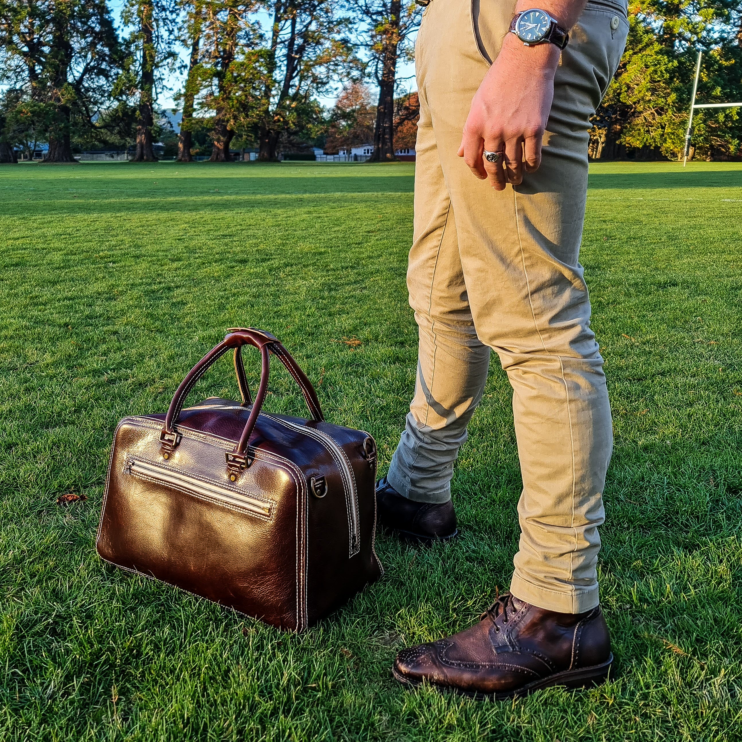 A lifestyle shot showing the No. 1963 VOYAGER Travel Bag in a glossy Tuscan Brown leather resting on bright green grass next to a person. The bag is angled to display its front zippered pocket, side vertical zipper, brass D-rings, and prominent white hand-stitching. The person standing beside it is visible from the waist down, styled in tan pants and brown leather brogue boots, set against a large park field with tall trees and goalposts illuminated by warm golden-hour sunlight.