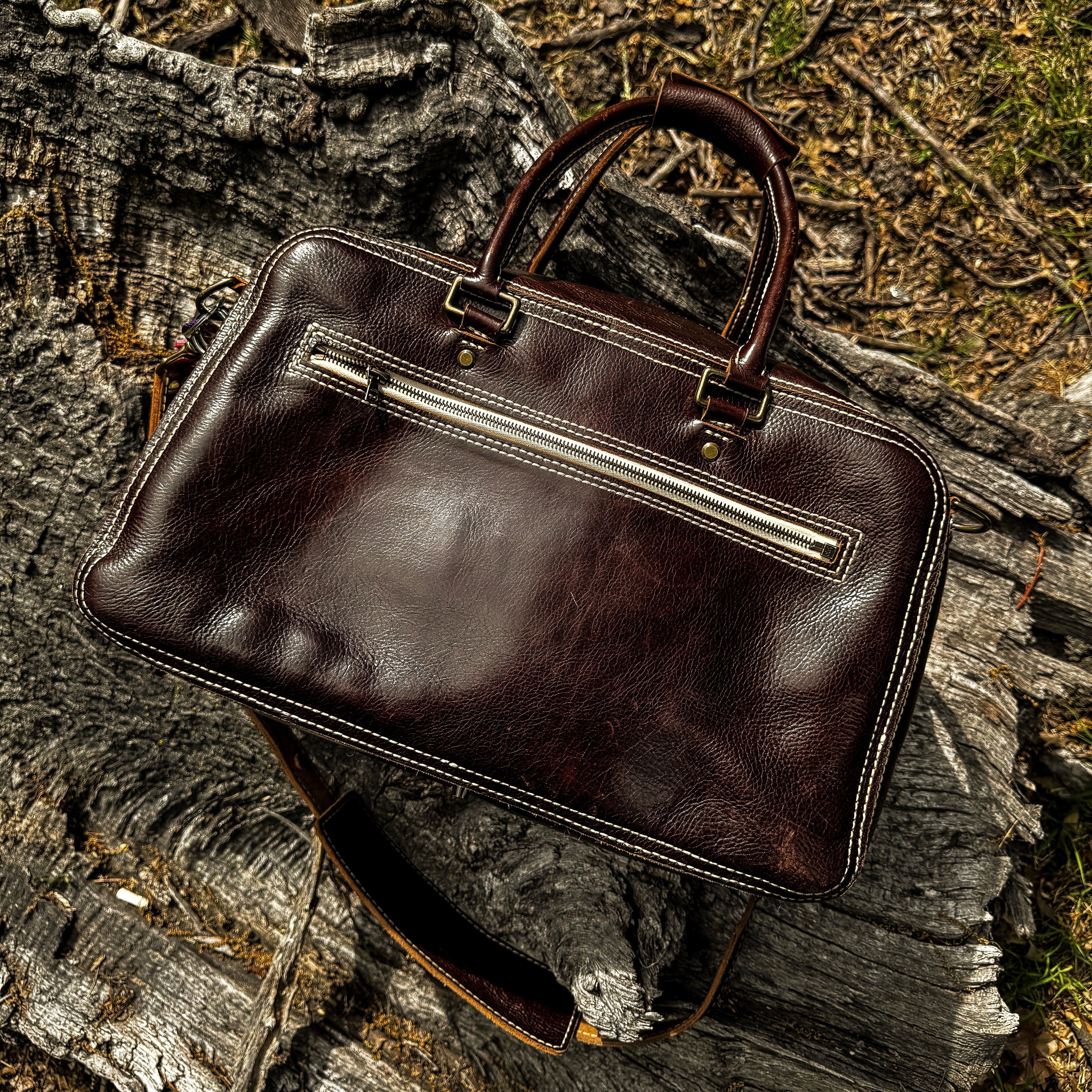 A top-down, angled lifestyle shot of the No. 1963 VOYAGER Travel Bag in a glossy Tuscan Brown leather, resting on a highly textured, weathered tree trunk. The image highlights the bag's prominent white hand-stitching, horizontal front zippered pocket, brass hardware, rolled top handles, and an attached shoulder strap draped beneath it, set against a natural outdoor background of rough wood and earth.