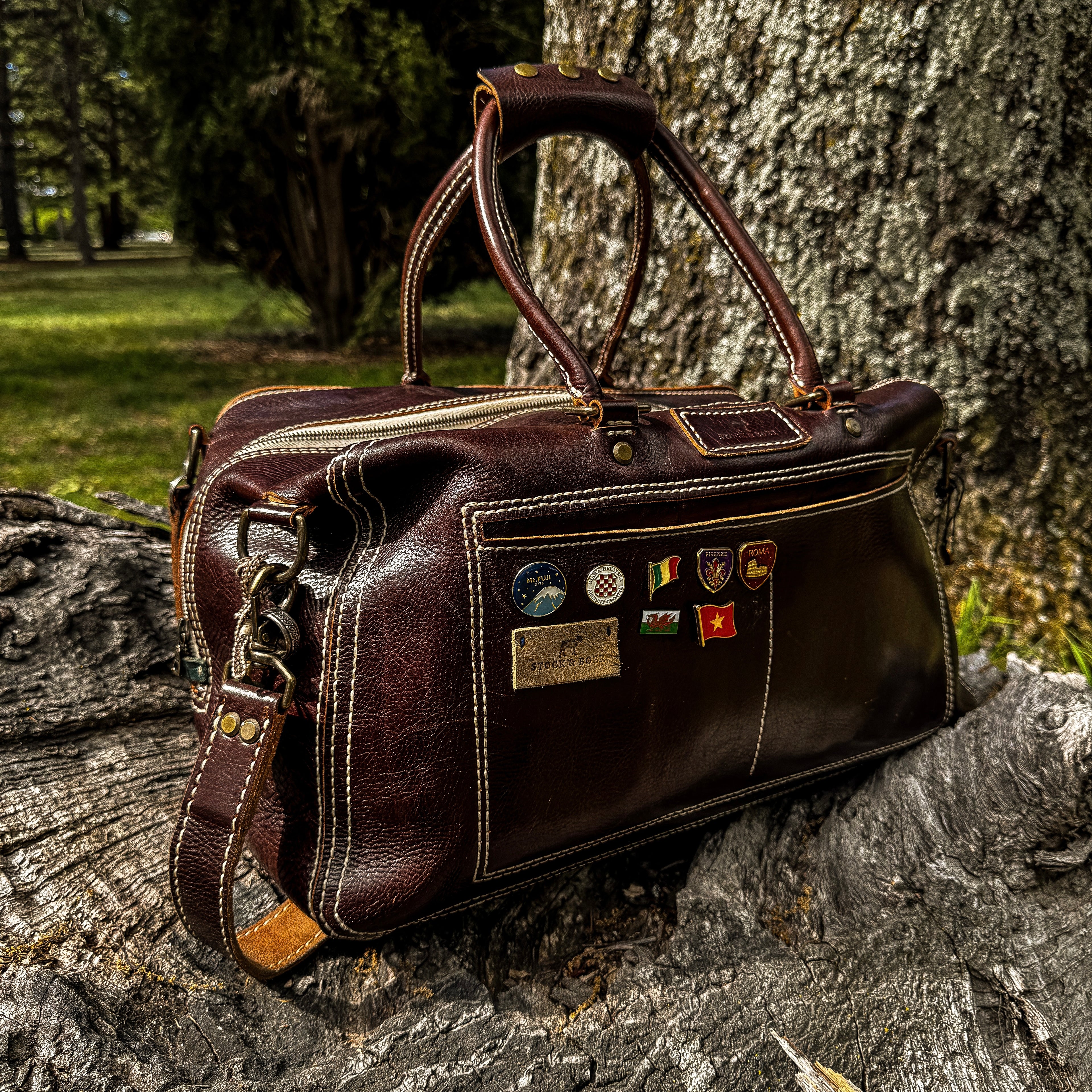 An angled lifestyle shot of the No. 1963 VOYAGER Travel Bag in a rich Tuscan Brown leather, resting on a heavily textured, weathered tree trunk. The bag features prominent white hand-stitching, brass hardware, rolled top handles, and an attached shoulder strap. The large front slip pocket is uniquely decorated with several colorful travel enamel pins and a small rectangular leather logo patch, set against a softly blurred background of a park with green grass and trees.