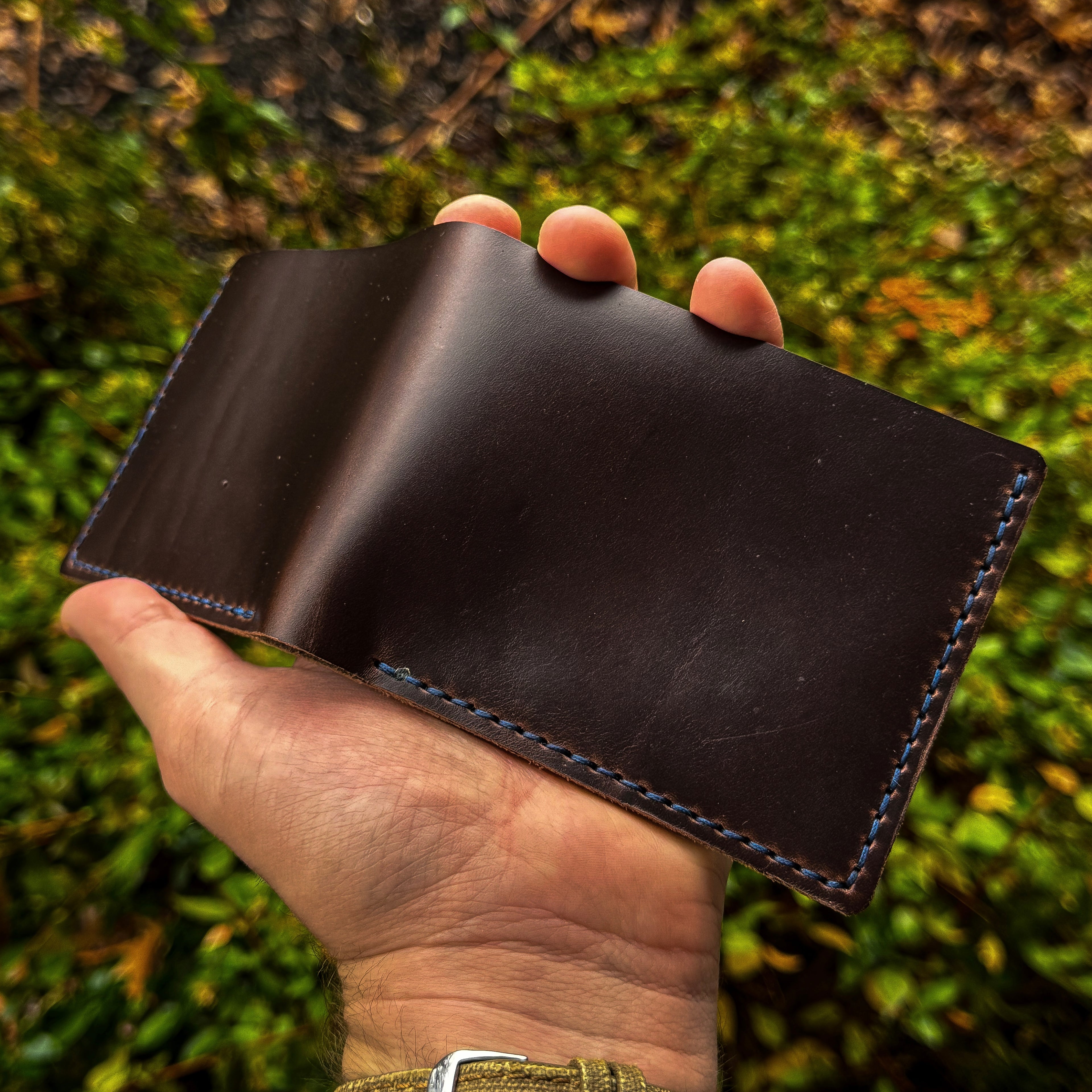 Hand holding an open No. 2 Frontiersman Leather Bifold Wallet handcrafted from Brown Chromexcel leather, featuring custom blue hand-stitching against a natural outdoor backdrop.