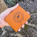 A close-up view of a hand presenting the closed front of the No. 2 Frontiersman Leather Bifold Wallet. Crafted from authentic Horween MLB Baseball Leather, the wallet showcases its signature deep orange-yellow color and rich texture. Contrast cream hand-stitching highlights the edges. A detailed circular Stock & Boer maker's mark is centrally debossed into the hide, displaying the logo and 'Handmade in New Zealand' text. The rustic natural backdrop of a tree trunk and forest floor provides context.