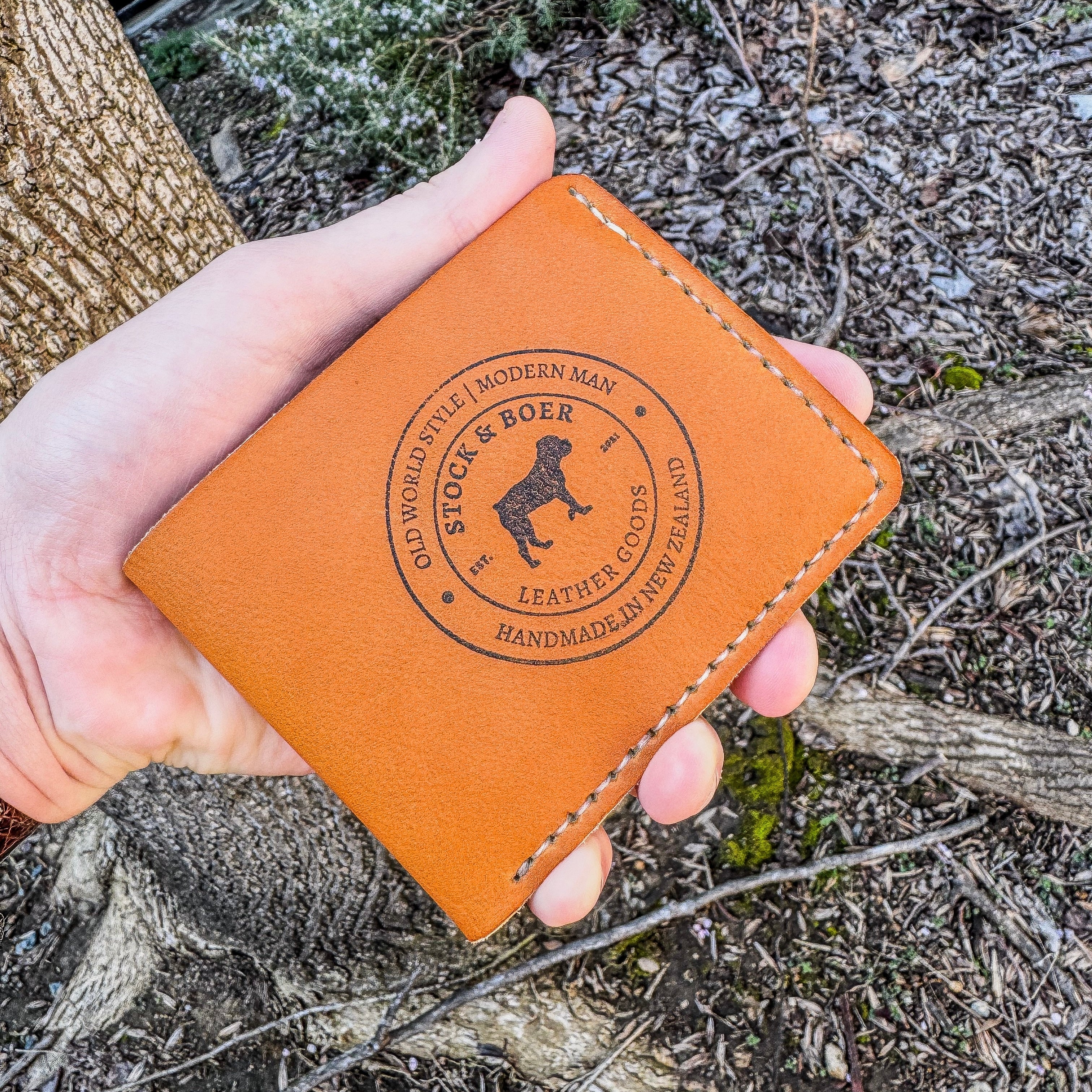 A close-up view of a hand presenting the closed front of the No. 2 Frontiersman Leather Bifold Wallet. Crafted from authentic Horween MLB Baseball Leather, the wallet showcases its signature deep orange-yellow color and rich texture. Contrast cream hand-stitching highlights the edges. A detailed circular Stock & Boer maker's mark is centrally debossed into the hide, displaying the logo and 'Handmade in New Zealand' text. The rustic natural backdrop of a tree trunk and forest floor provides context.