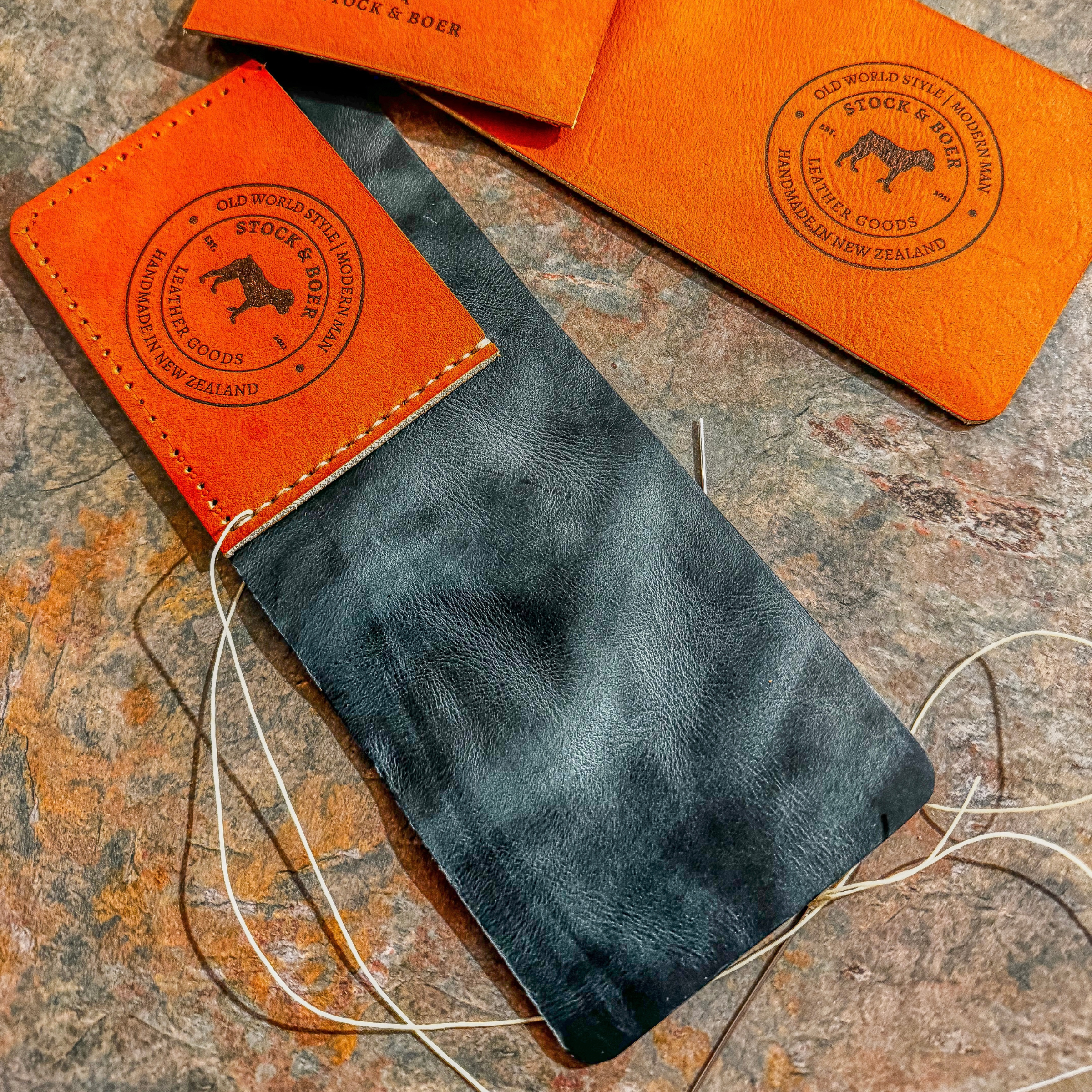 A behind-the-scenes look at the artisanal construction of the No. 2 Frontiersman Leather Bifold Wallet. The image displays the vibrant orange Horween MLB Baseball leather exterior, complete with the prominently debossed Stock & Boer maker's mark, actively being hand-stitched to a contrasting, richly grained black leather interior panel. Heavy waxed thread and needles rest on a rugged, textured workbench, emphasizing the authentic, meticulous handcrafted process.
