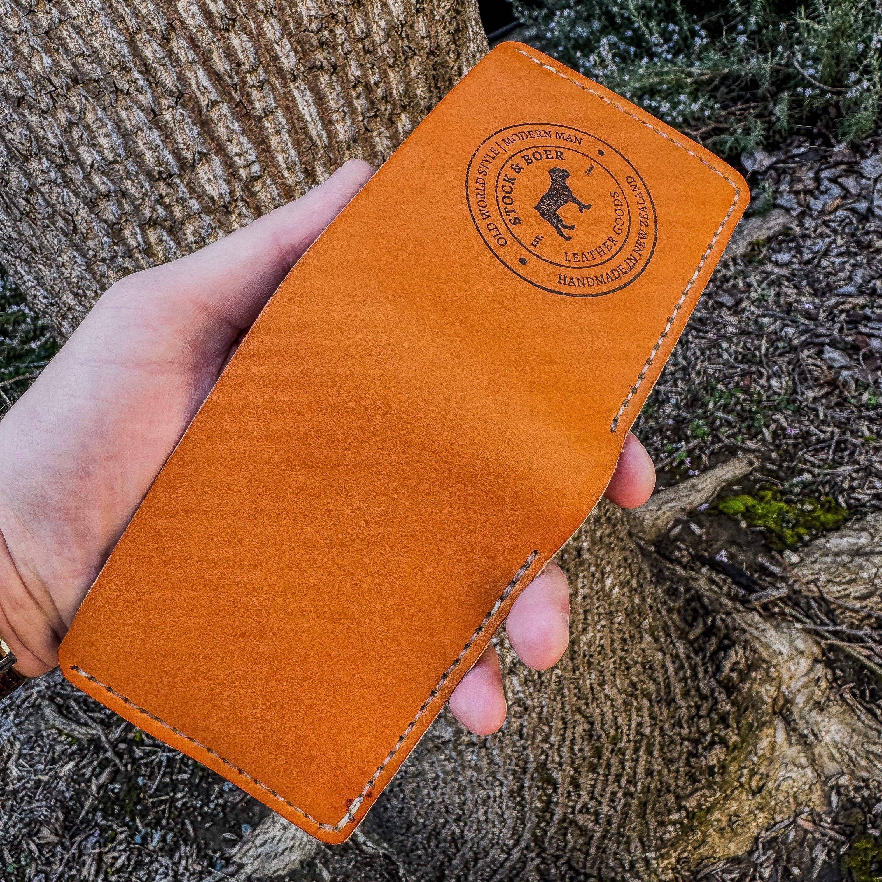 A handheld view of the No. 2 Frontiersman Leather Bifold Wallet laid completely open and flat, revealing the full, uninterrupted exterior crafted from premium Horween MLB Baseball leather. The image highlights the distinctive, vibrant orange tone and smooth grain of the hide, bordered by precise cream-colored perimeter hand-stitching. The right panel features the sharply ink stamped circular Stock & Boer maker's mark, all set against a deeply textured tree bark and forest floor background.