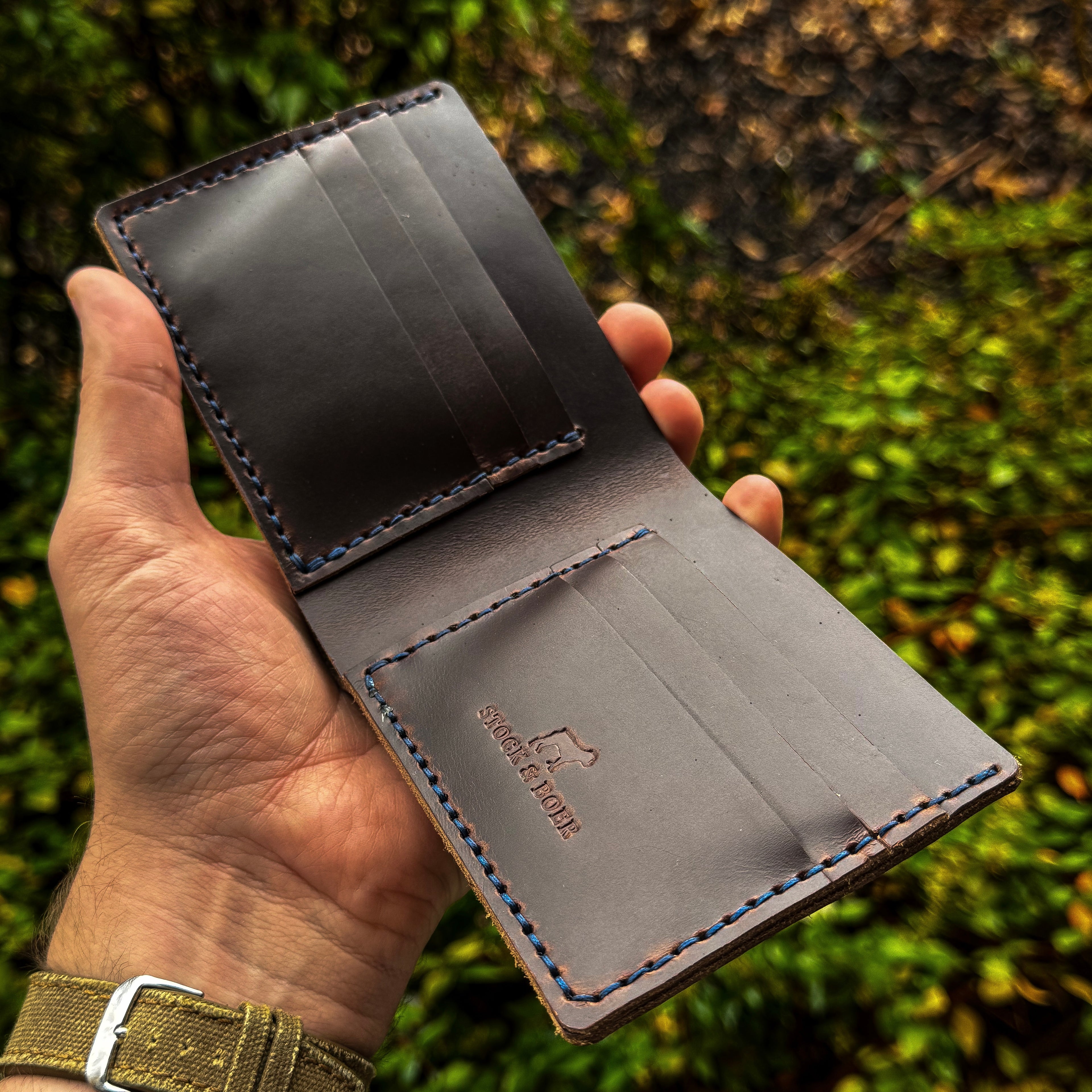 Interior view of the No. 2 Frontiersman Leather Bifold Wallet held in hand, handcrafted from Brown Chromexcel with custom blue stitching. Features a multi-card layout and the Stock & Boer embossed logo against a natural woodland background.