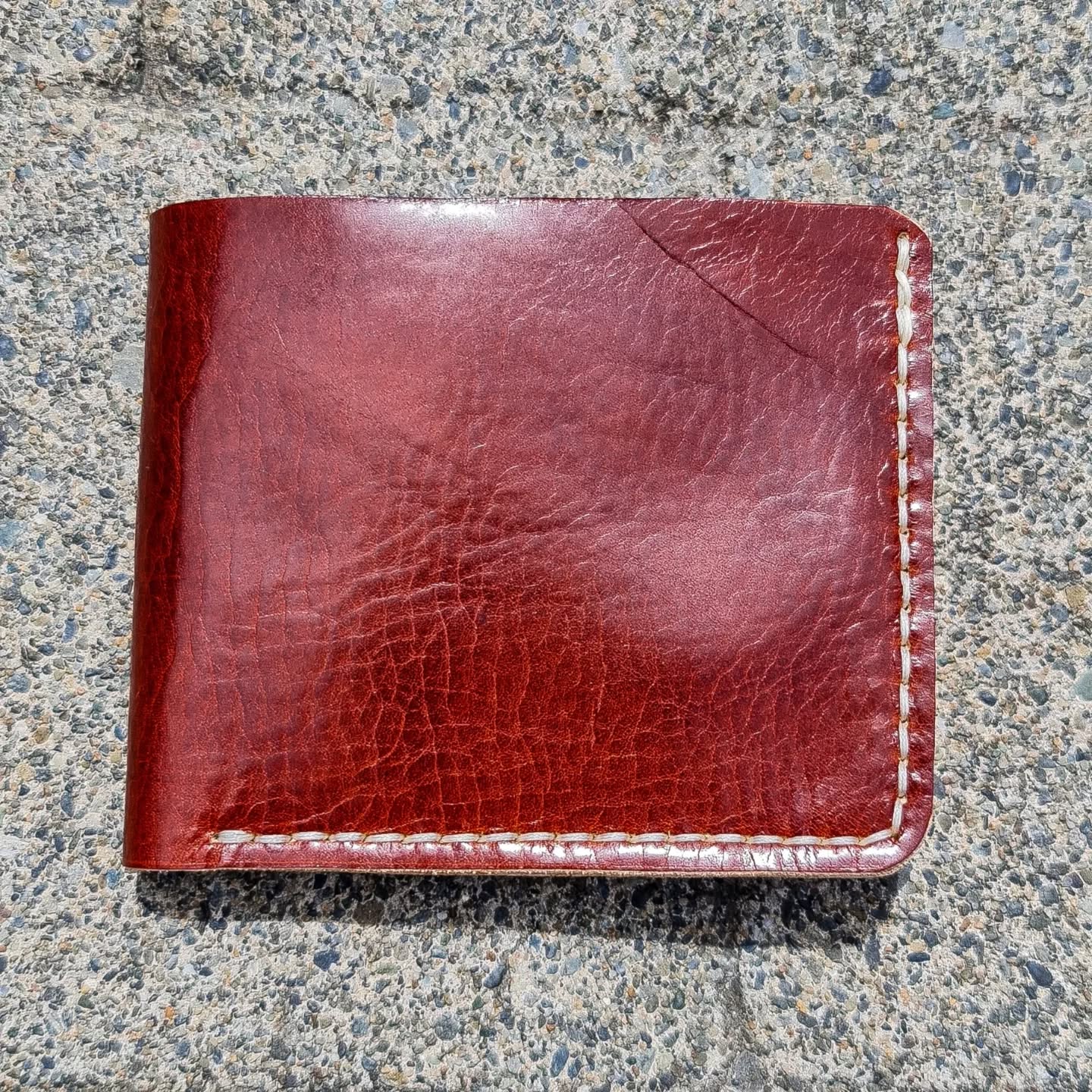 A closed No. 2 FRONTIERSMAN Leather Bifold Wallet in a rich reddish-brown leather, lying flat on a textured concrete surface. The exterior showcases a natural, slightly glossy grain texture and features contrasting white perimeter stitching.