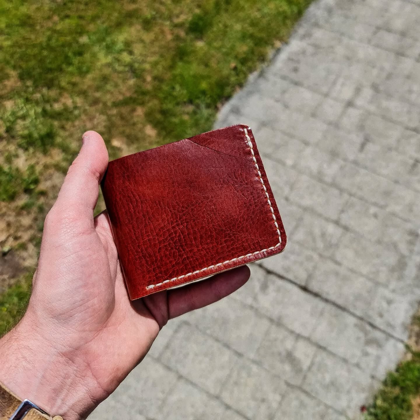 A hand holding a closed No. 2 FRONTIERSMAN Leather Bifold Wallet in a rich cognac-colored leather. The exterior showcases a natural grain texture and contrasting white perimeter stitching, set against a blurred outdoor background of grass and a paved path.