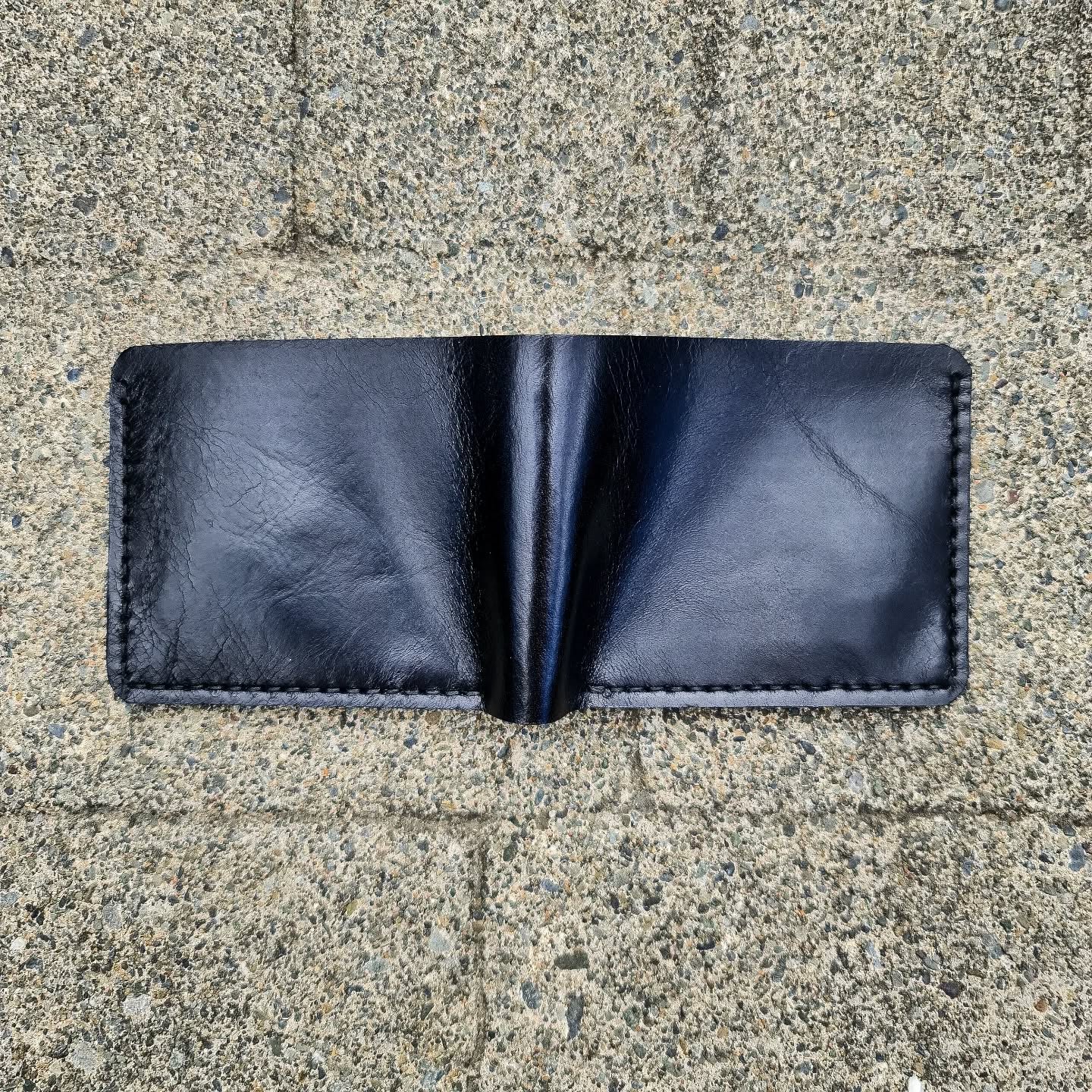 A fully opened black No. 2 FRONTIERSMAN Leather Bifold Wallet, displaying its smooth La Bretagna leather exterior, central fold, and perimeter stitching, laid flat against a textured concrete background.