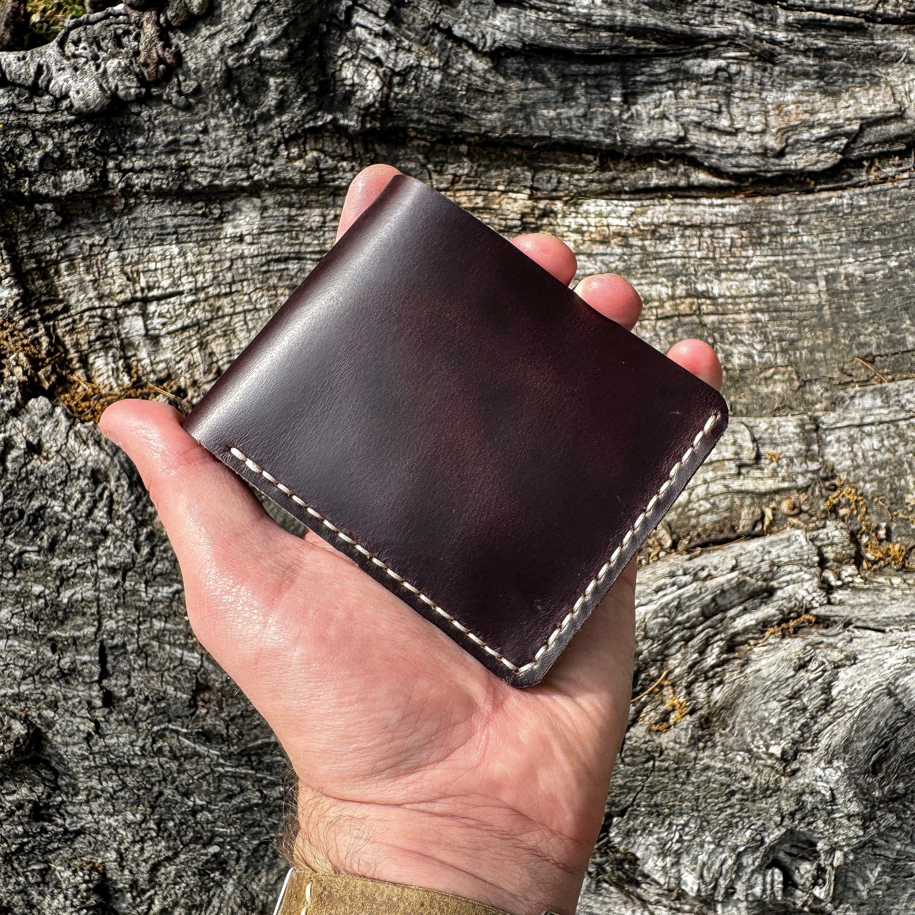 A hand holding the No. 2 FRONTIERSMAN Leather Bifold Wallet, crafted from burgundy Horween Chromexcel leather, against a textured, rugged bark background. The image showcases the wallet's minimalist bifold design, the deep, rich tones of the burgundy leather, and the precise contrast stitching.