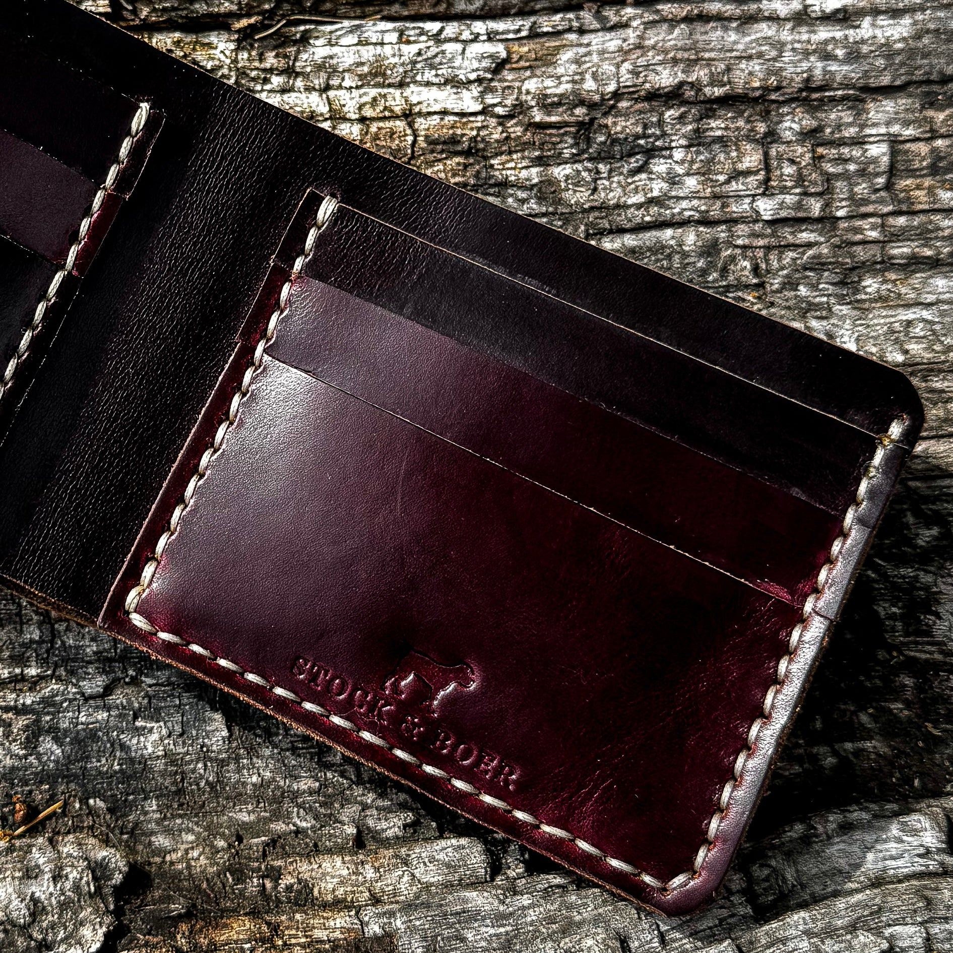 A detailed, interior view of the open No. 2 FRONTIERSMAN Leather Bifold Wallet, crafted from burgundy Horween Chromexcel leather. The image highlights the internal card slots, precise contrast stitching, and the stamped "STOCK & BOER" logo, all set against a textured, rugged bark background.