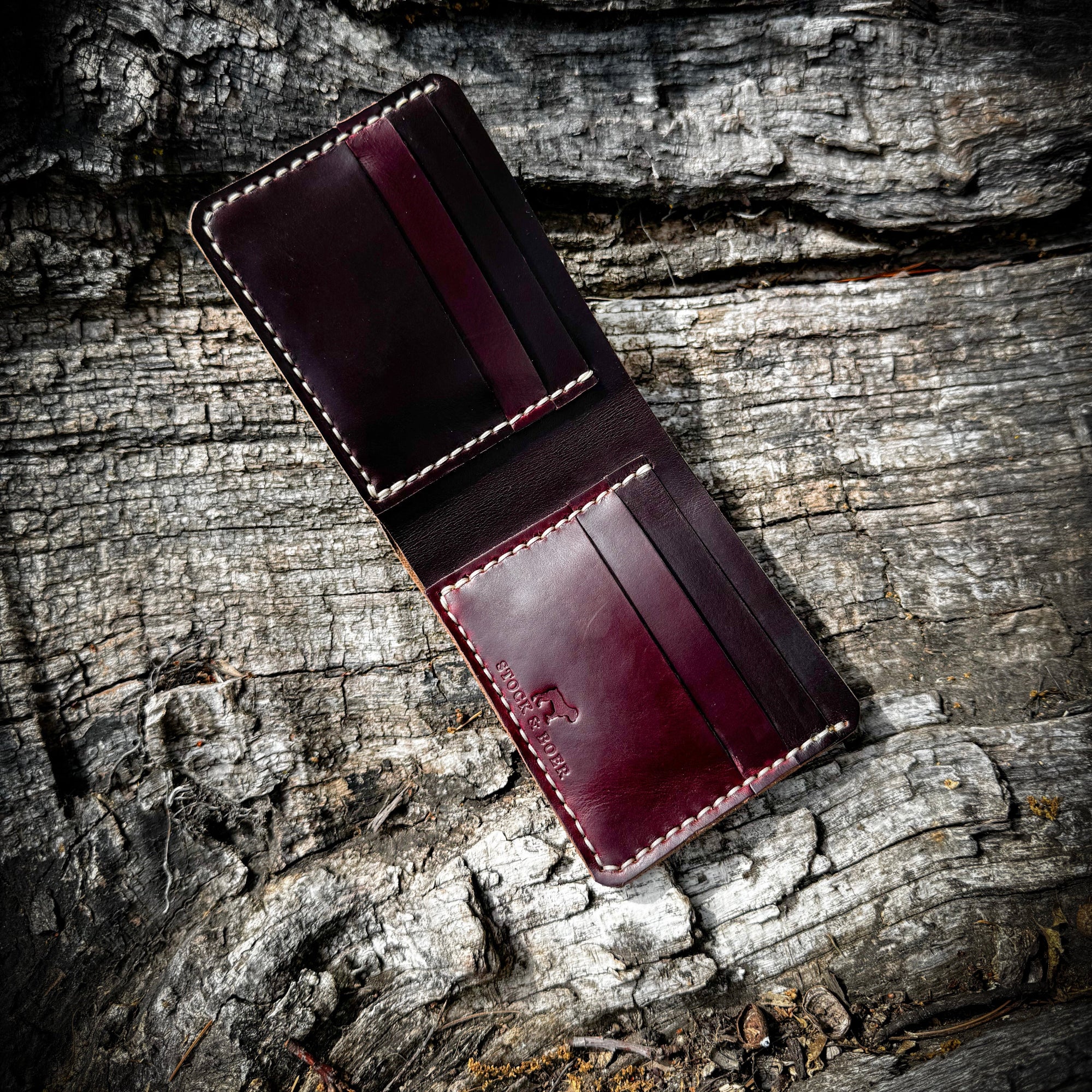 A top-down, flat-lay view of the open No. 2 FRONTIERSMAN Leather Bifold Wallet, crafted from burgundy Horween Chromexcel leather. The image showcases the wallet's internal card slots, precise contrast stitching, and the stamped "STOCK & BOER" logo, all resting on a textured, rugged bark background.