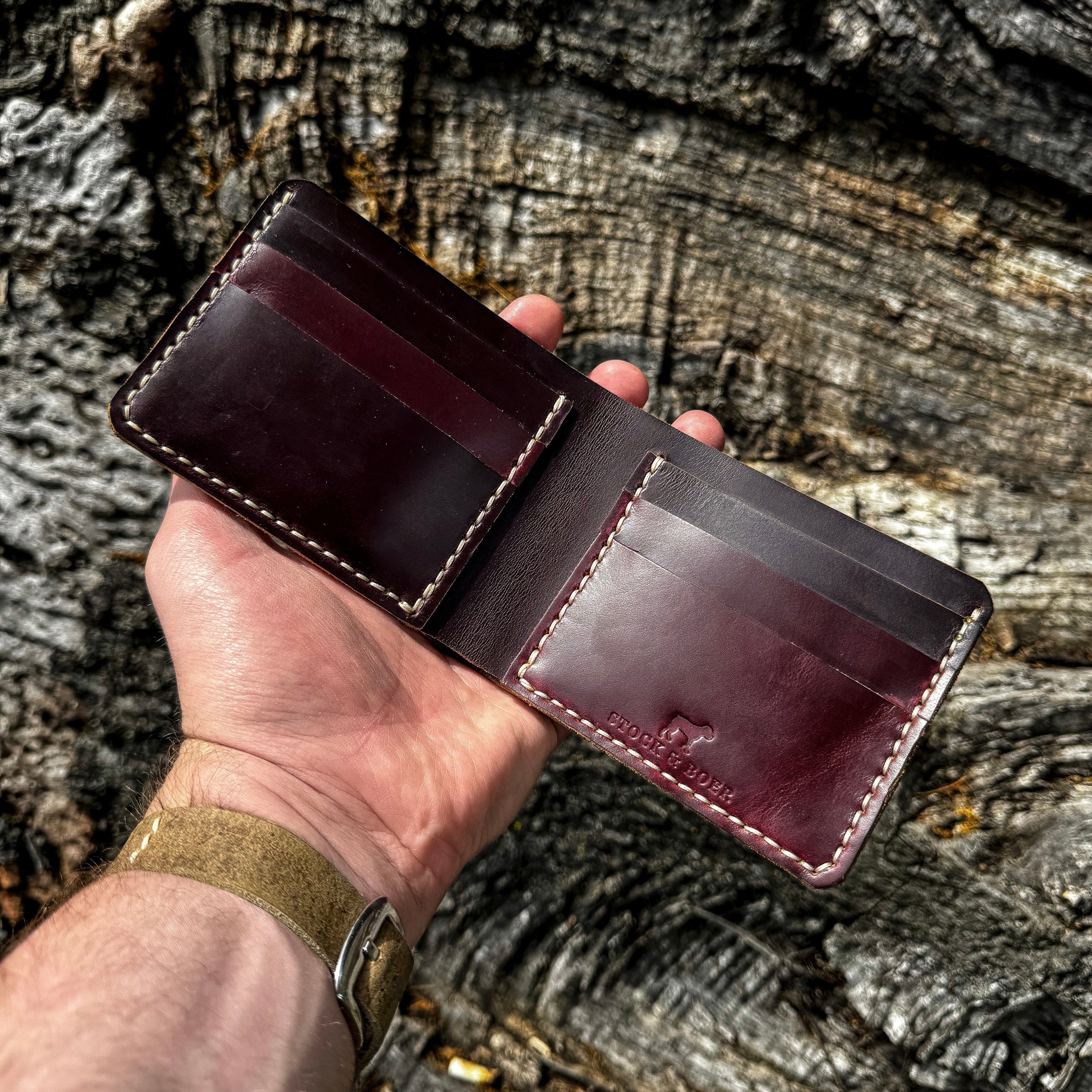 A hand holding the open No. 2 FRONTIERSMAN Leather Bifold Wallet, crafted from burgundy Horween Chromexcel leather, against a textured, rugged bark background. The image highlights the internal card slots, precise contrast stitching, and the stamped "STOCK & BOER" logo.