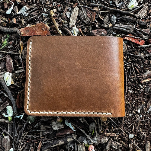 A closed, back-view of the No. 2 FRONTIERSMAN Leather Bifold Wallet, crafted from natural Horween Chromexcel leather. The wallet is resting on a ground covered in mulch and white flower petals, showcasing its clean, minimalist design, rich texture, and precise contrast stitching.
