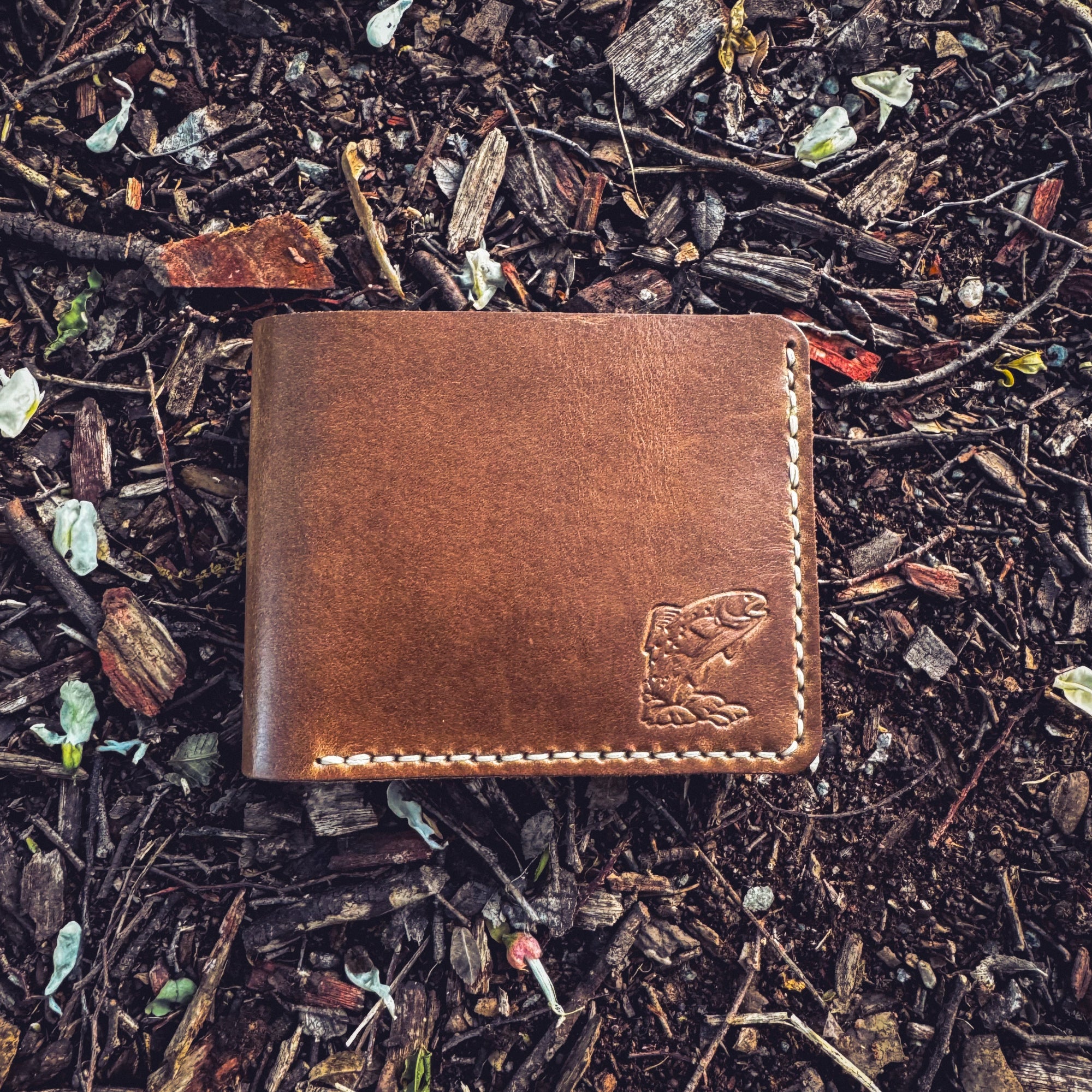 A flat-lay view of the No. 2 FRONTIERSMAN Leather Bifold Wallet in natural Horween Chromexcel leather, featuring custom trout embossing. The wallet is positioned on a ground covered in mulch and white flower petals, showcasing the debossed fish design, the rich texture of the natural leather, and the precise contrast stitching.
